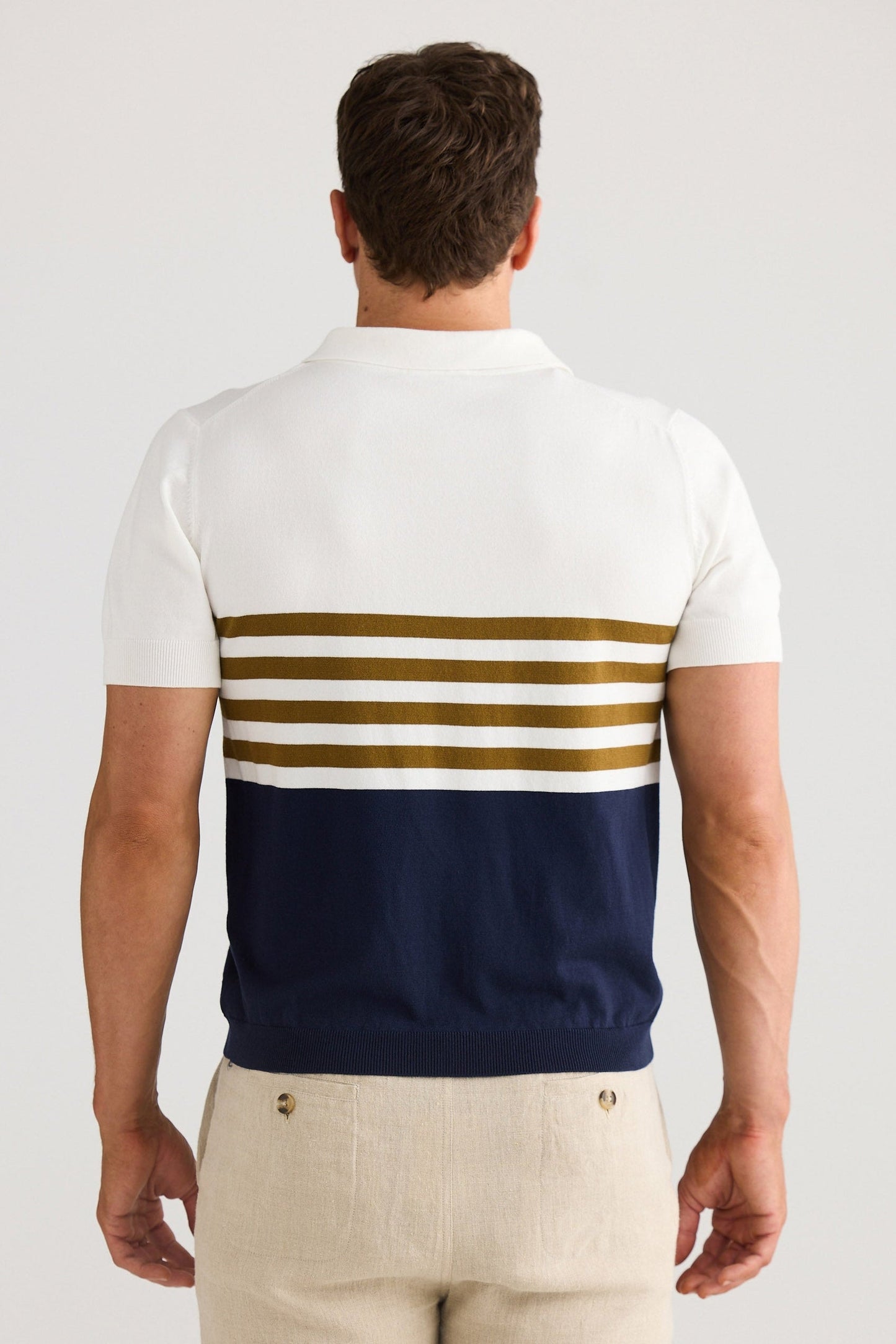 Man wearing a white polo shirt with blue and gold stripes and beige shorts on a light gray background