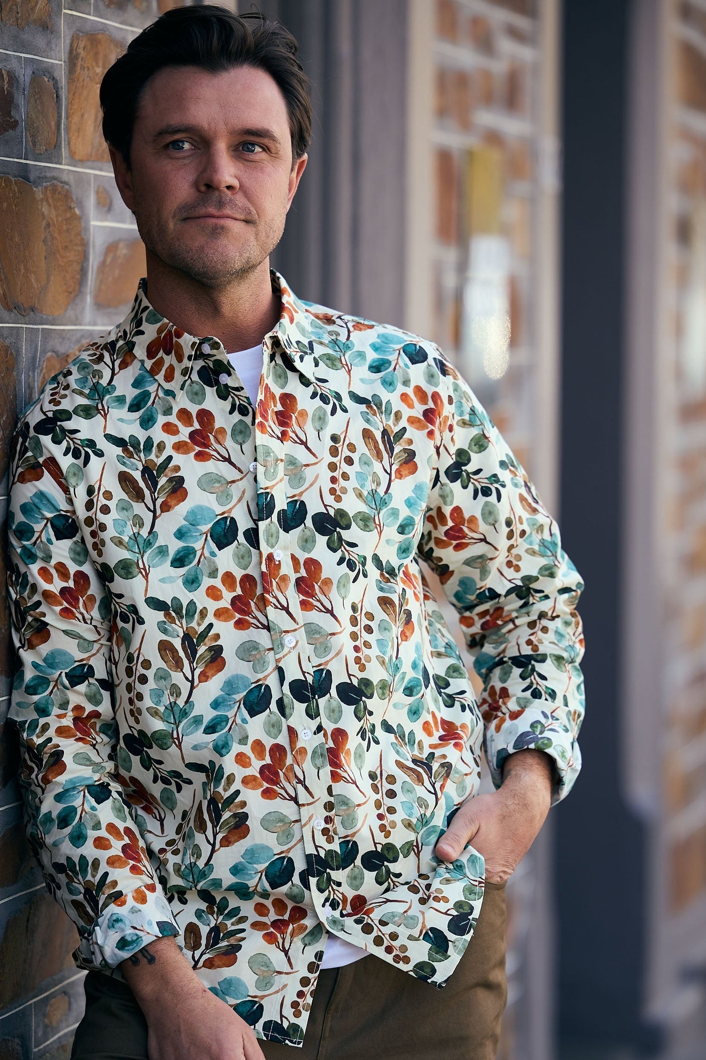 Man wearing a colorful floral shirt leaning against a brick wall.