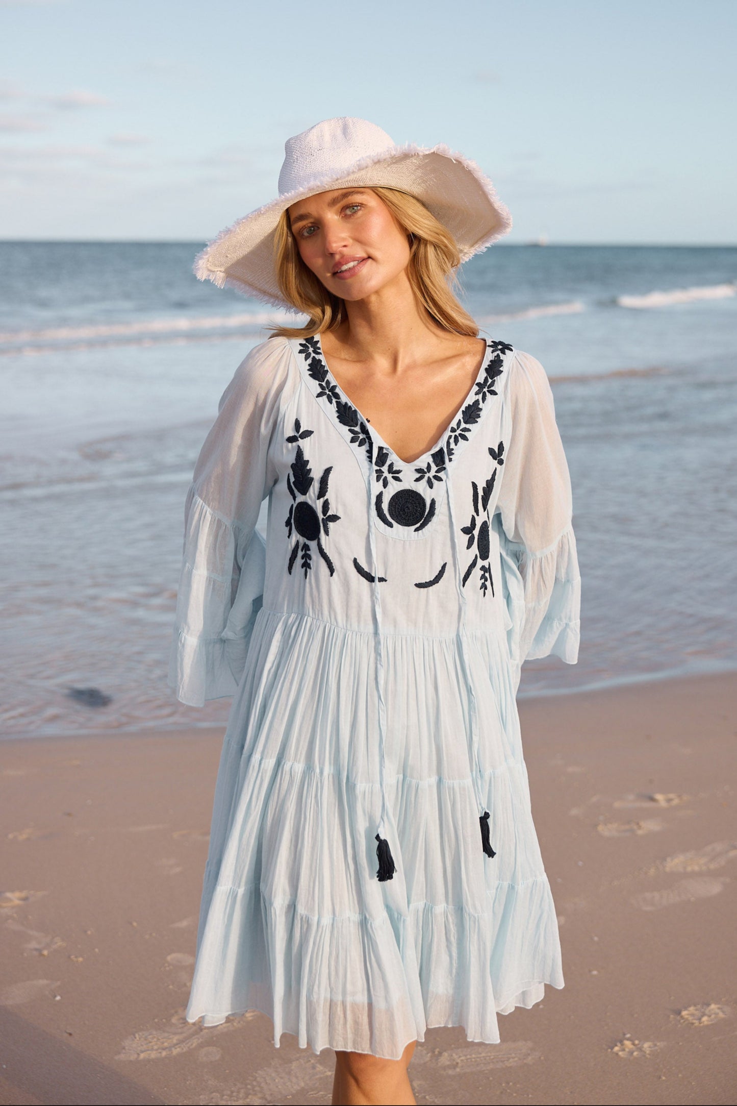 Woman in a light blue dress with navy embroidery and a wide-brimmed hat standing on a beach.