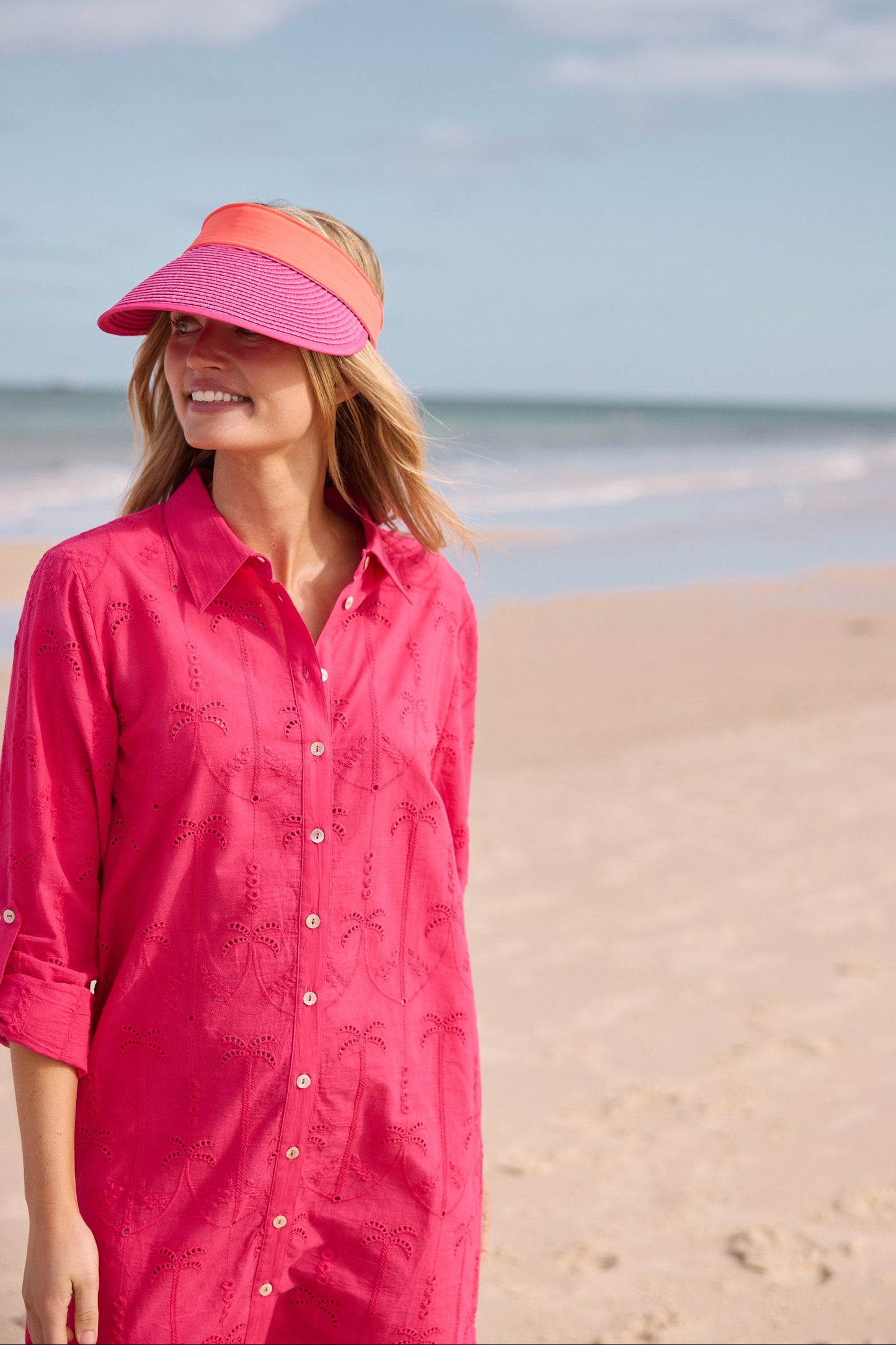 Woman in a pink shirt dress and visor standing on a sandy beach with ocean in the background