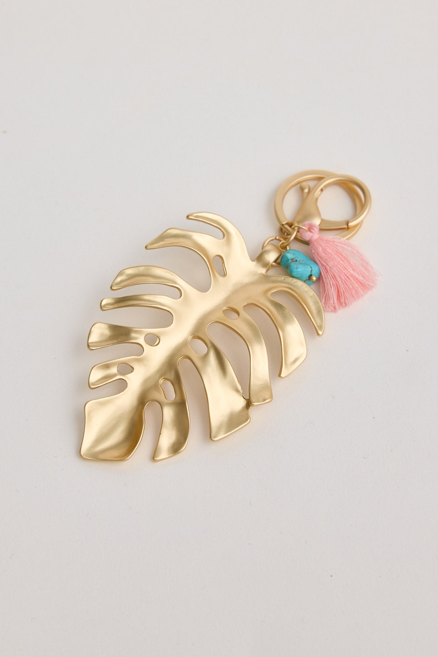 Gold leaf-shaped keychain with pink tassel and turquoise stone on a light gray background