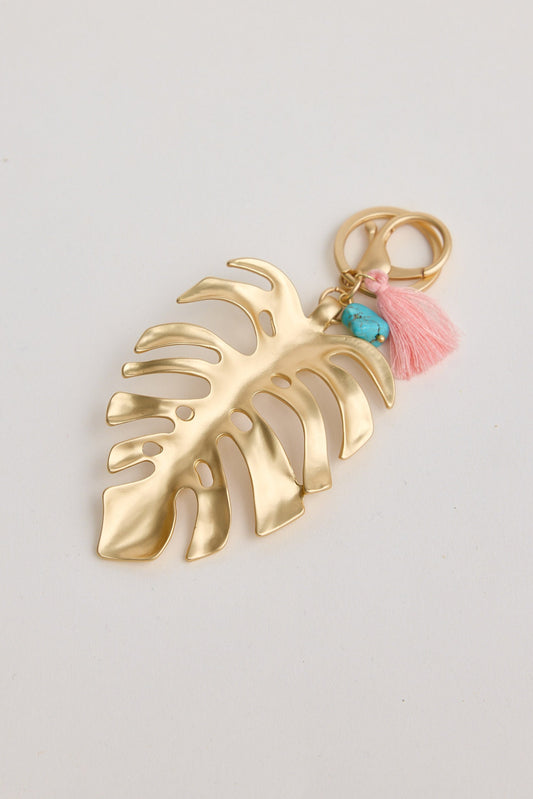 Gold leaf-shaped keychain with pink tassel and turquoise stone on a light gray background