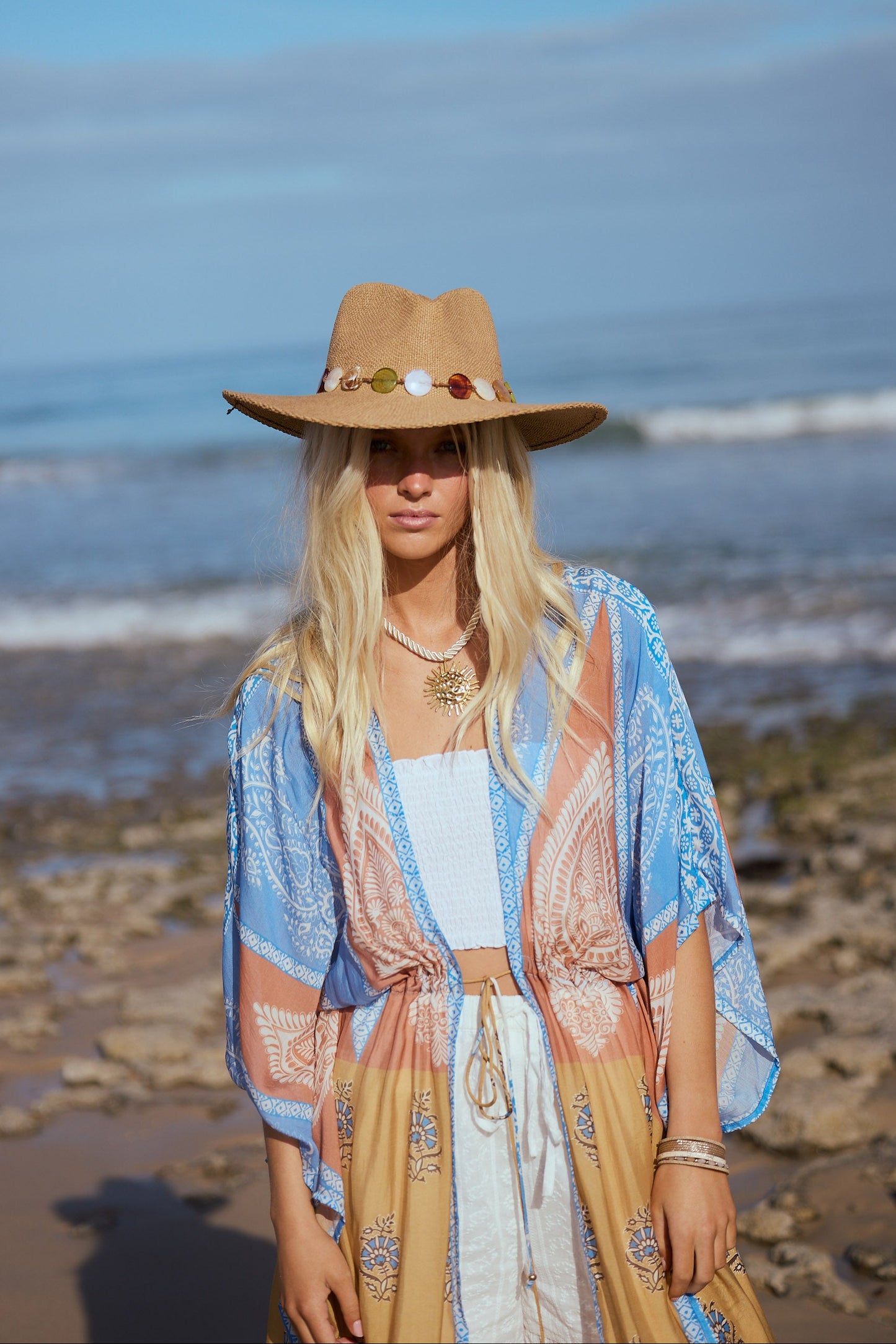 Woman in a hat and patterned outfit standing on a beach with ocean waves in the background