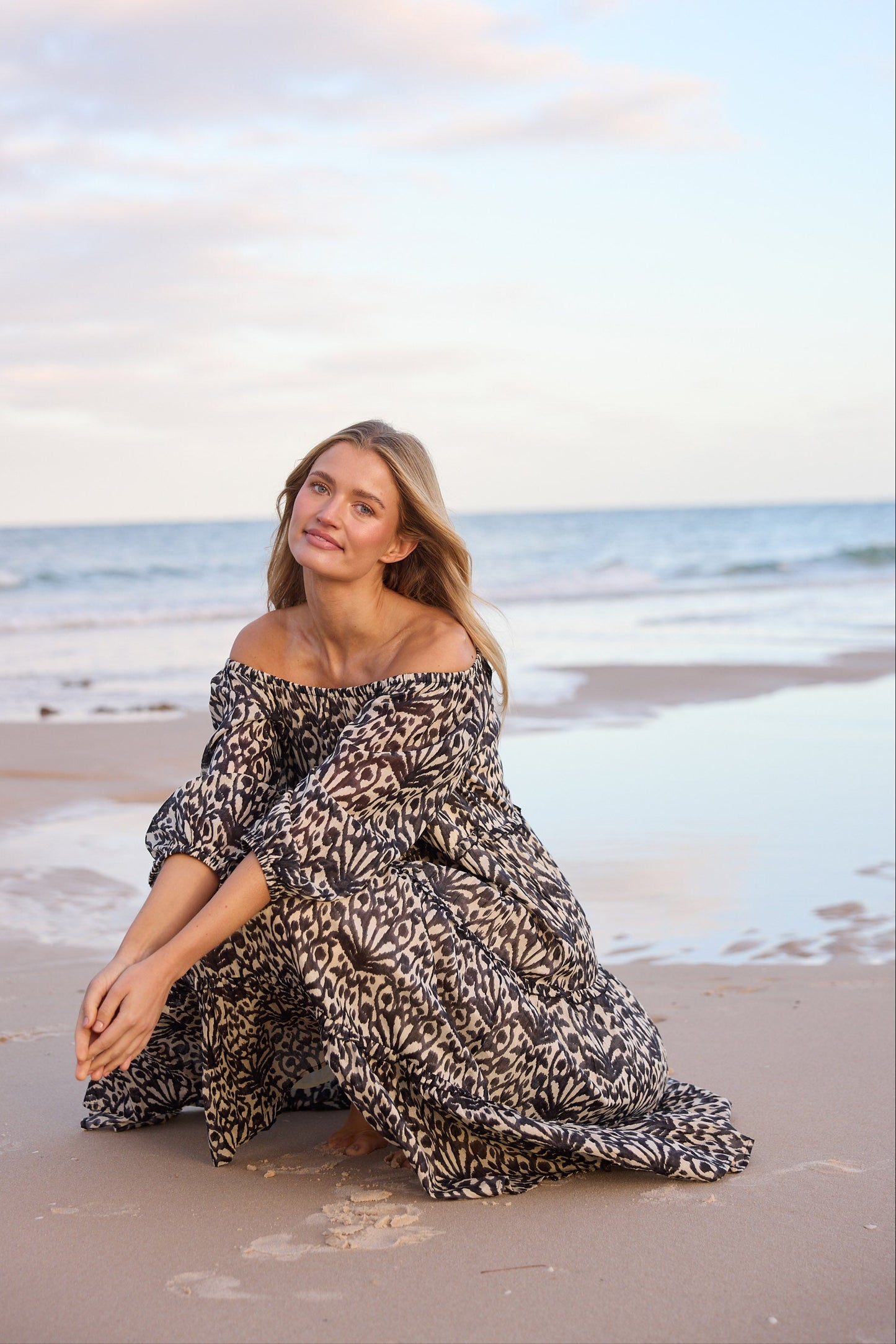 Woman in a patterned dress sitting on a beach with ocean and sky in the background