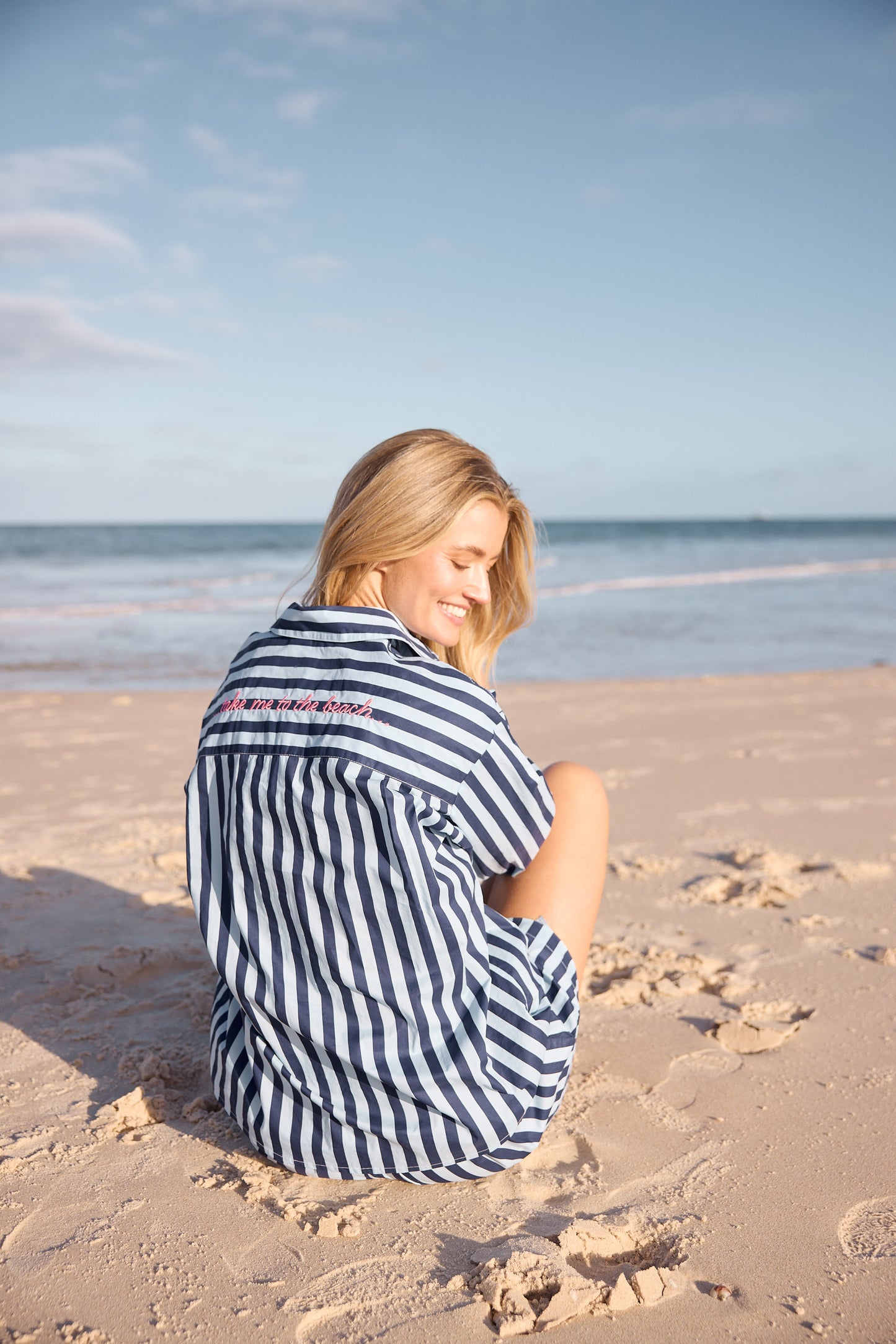 Woman sitting on a sandy beach wearing a striped shirt with ocean and sky in the background