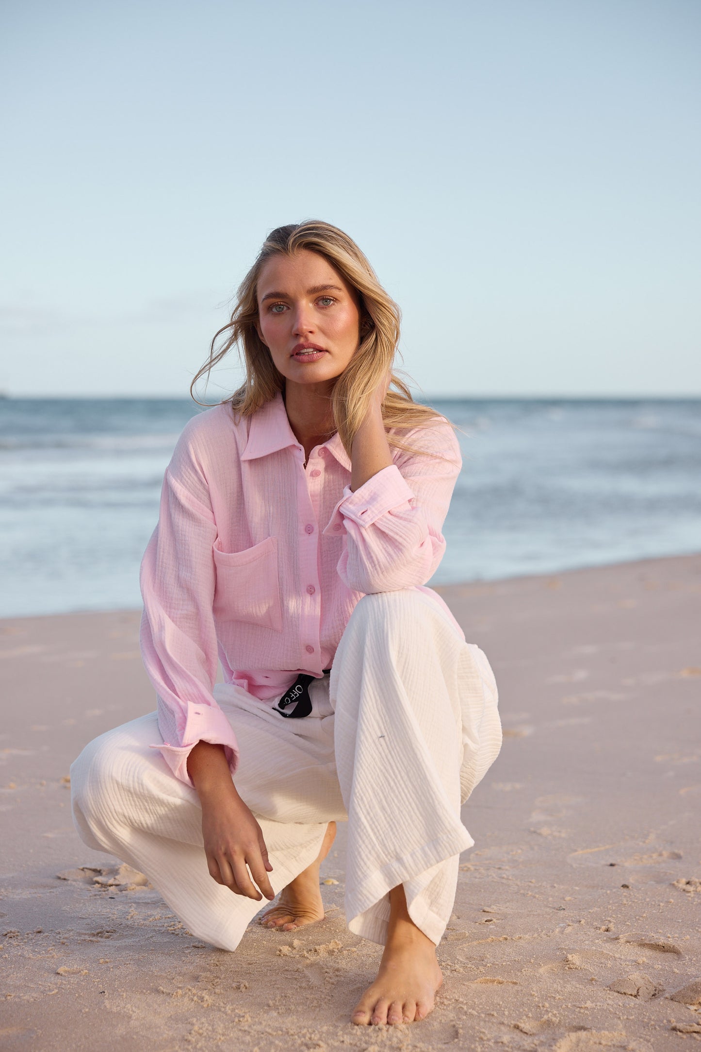 Woman in pink shirt and white pants sitting on a beach with ocean in the background