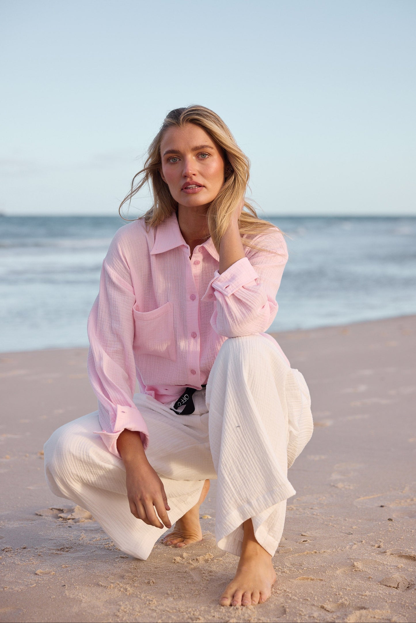 Woman in pink shirt and white pants sitting on a beach with ocean in the background