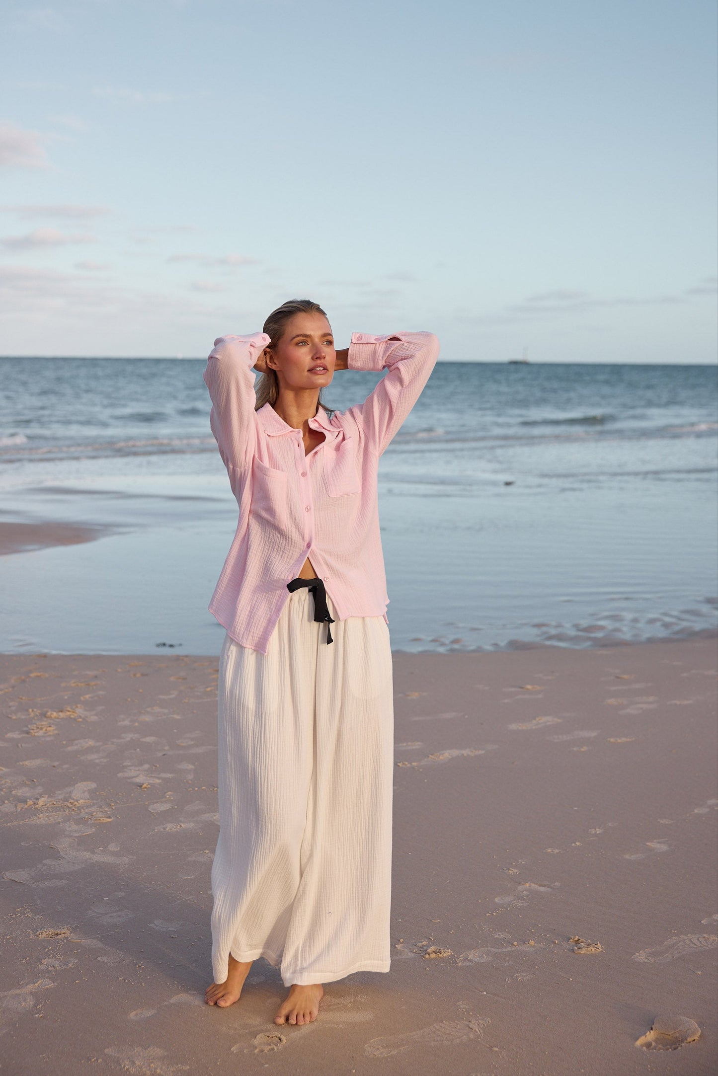 Woman in a pink shirt and white pants standing on a beach with ocean and sky in the background