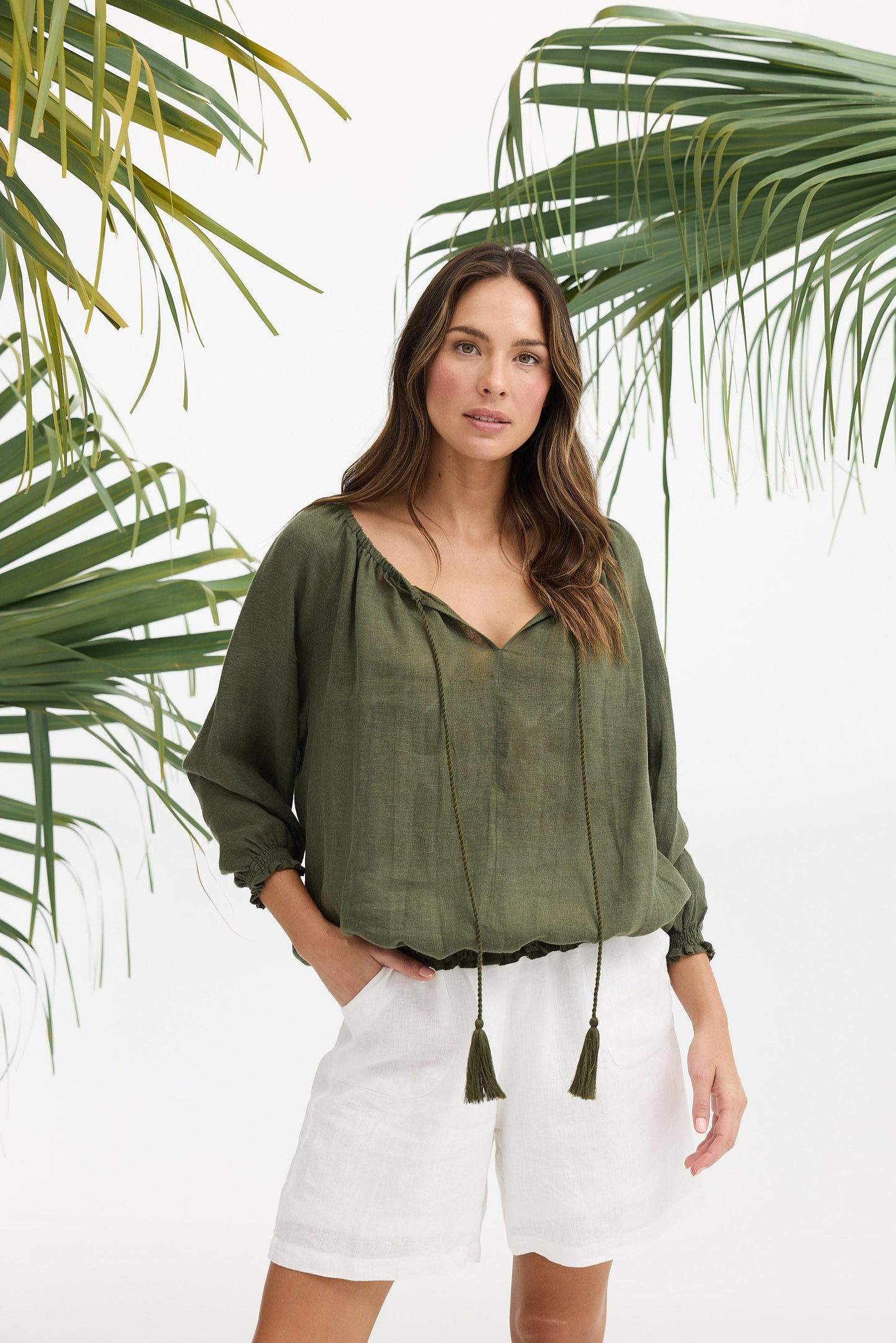 Woman wearing a green blouse and white shorts standing in front of palm leaves.