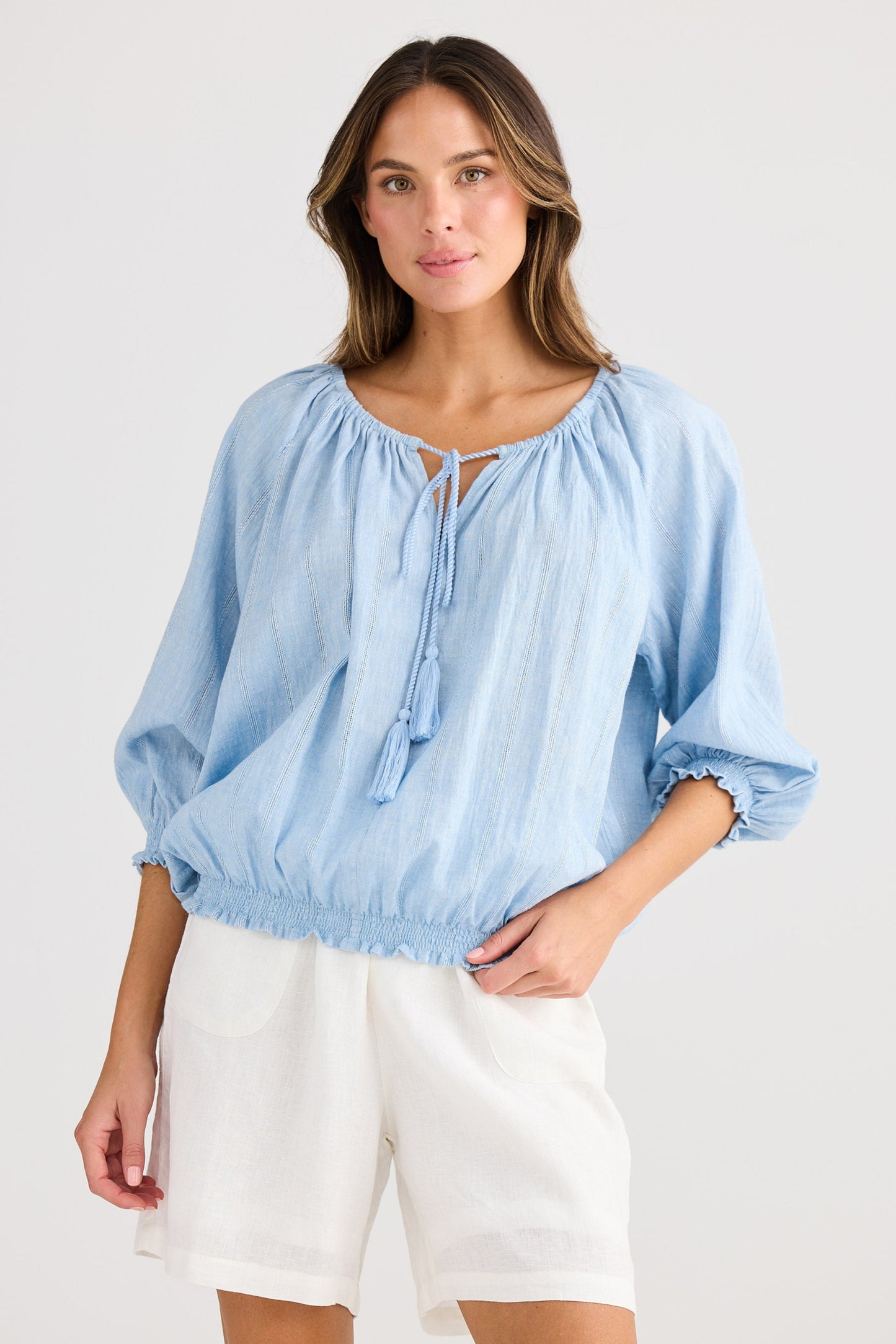 Woman wearing a light blue blouse with balloon sleeves and white shorts on a plain background
