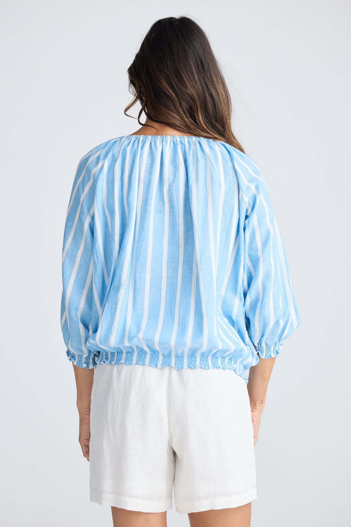 Woman wearing a light blue striped blouse and white shorts on a plain background