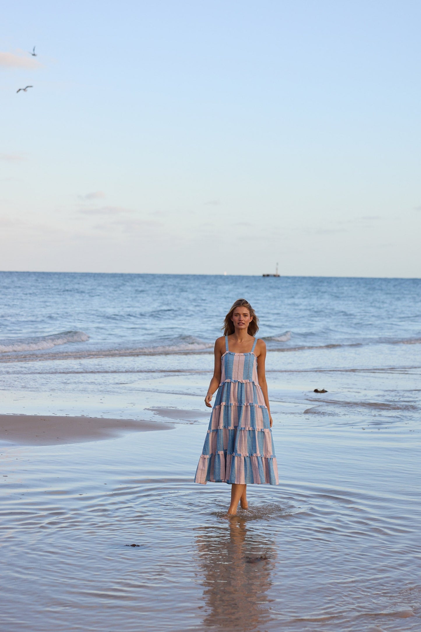 Woman in a striped dress standing in shallow water on a beach with a clear sky.