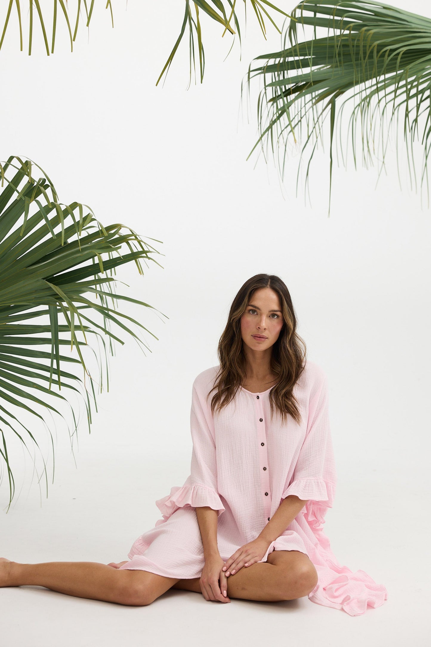 Woman in a pink dress sitting on a white background with palm leaves.