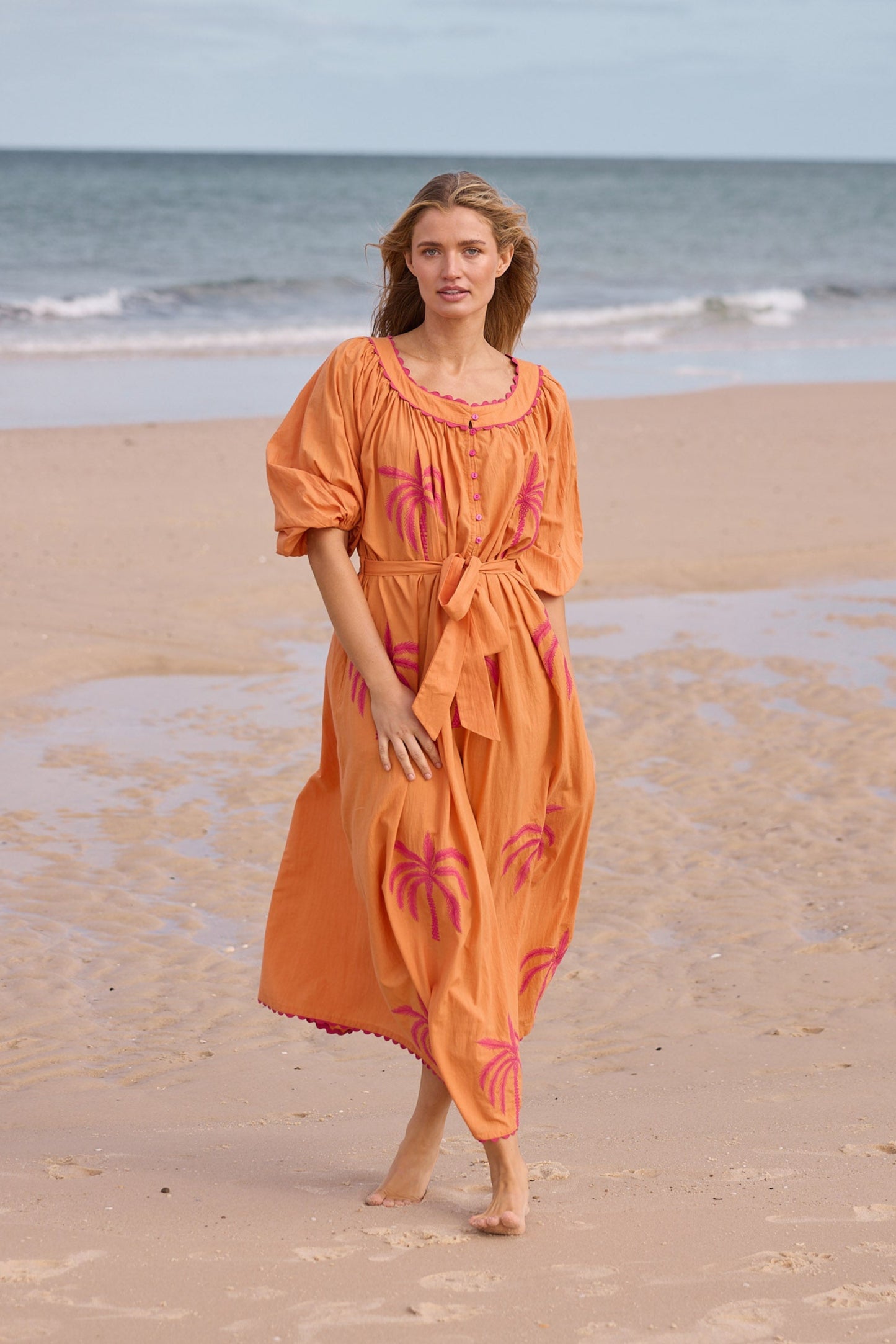 Woman in an orange dress with pink floral patterns walking on a beach.