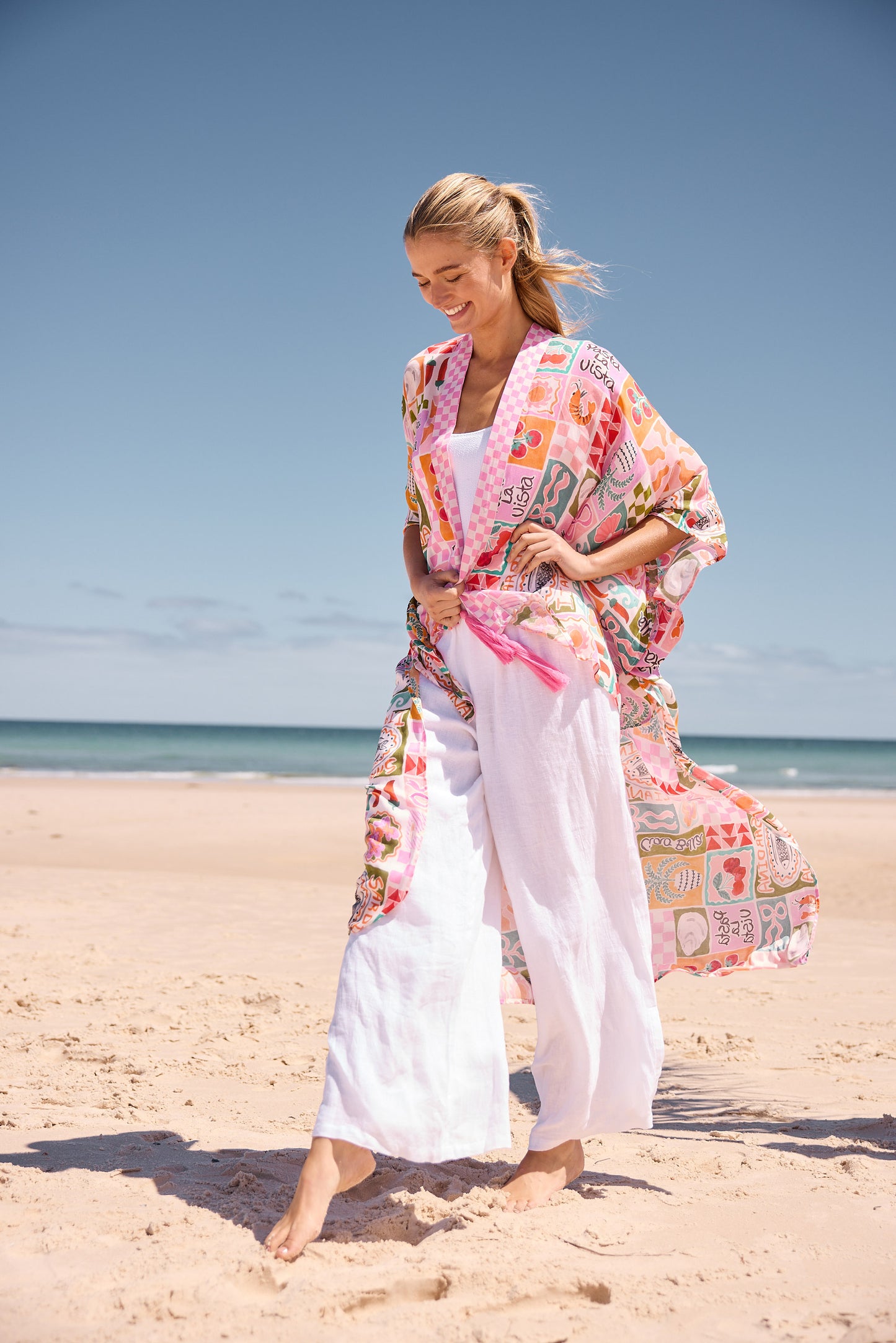 Woman in a colorful kimono and white pants standing on a sandy beach with blue sky.