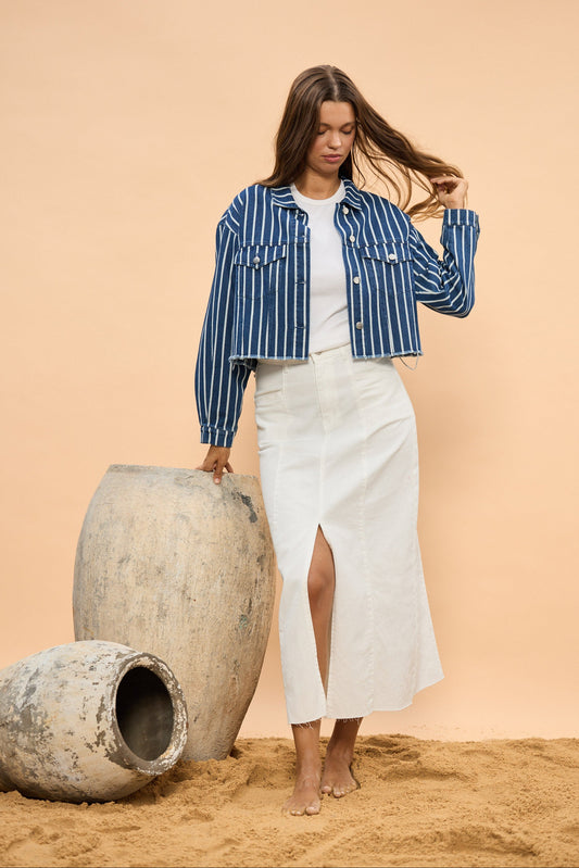 Woman in a blue striped shirt and white skirt standing next to large stone pots on a sandy surface.