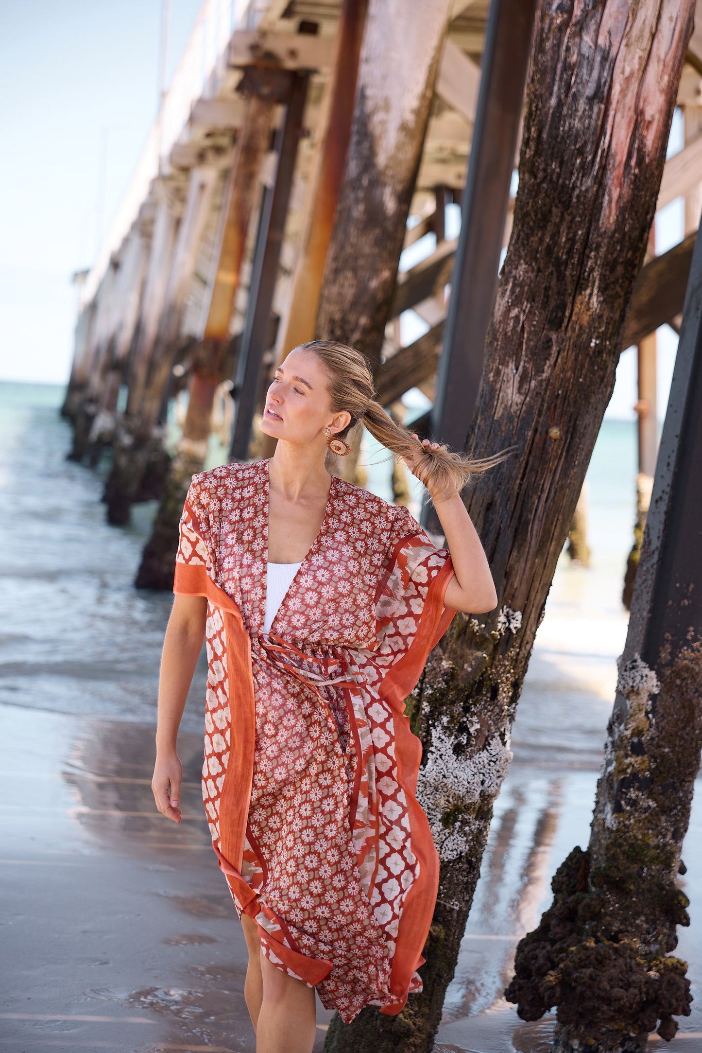 Woman in a orange and white patterned kaftan standing on a beach near a pier.