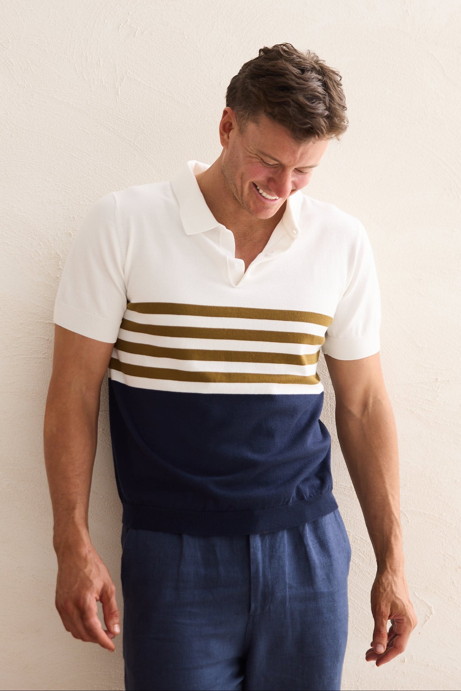 Man wearing a white polo shirt with navy and gold stripes and navy pants against a plain background
