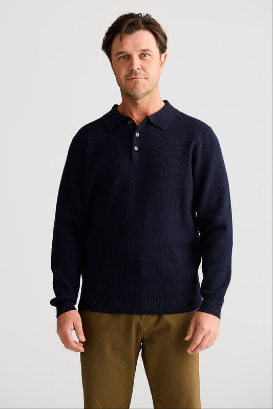 Man wearing a navy sweater over a collared shirt with brown pants on a light gray background