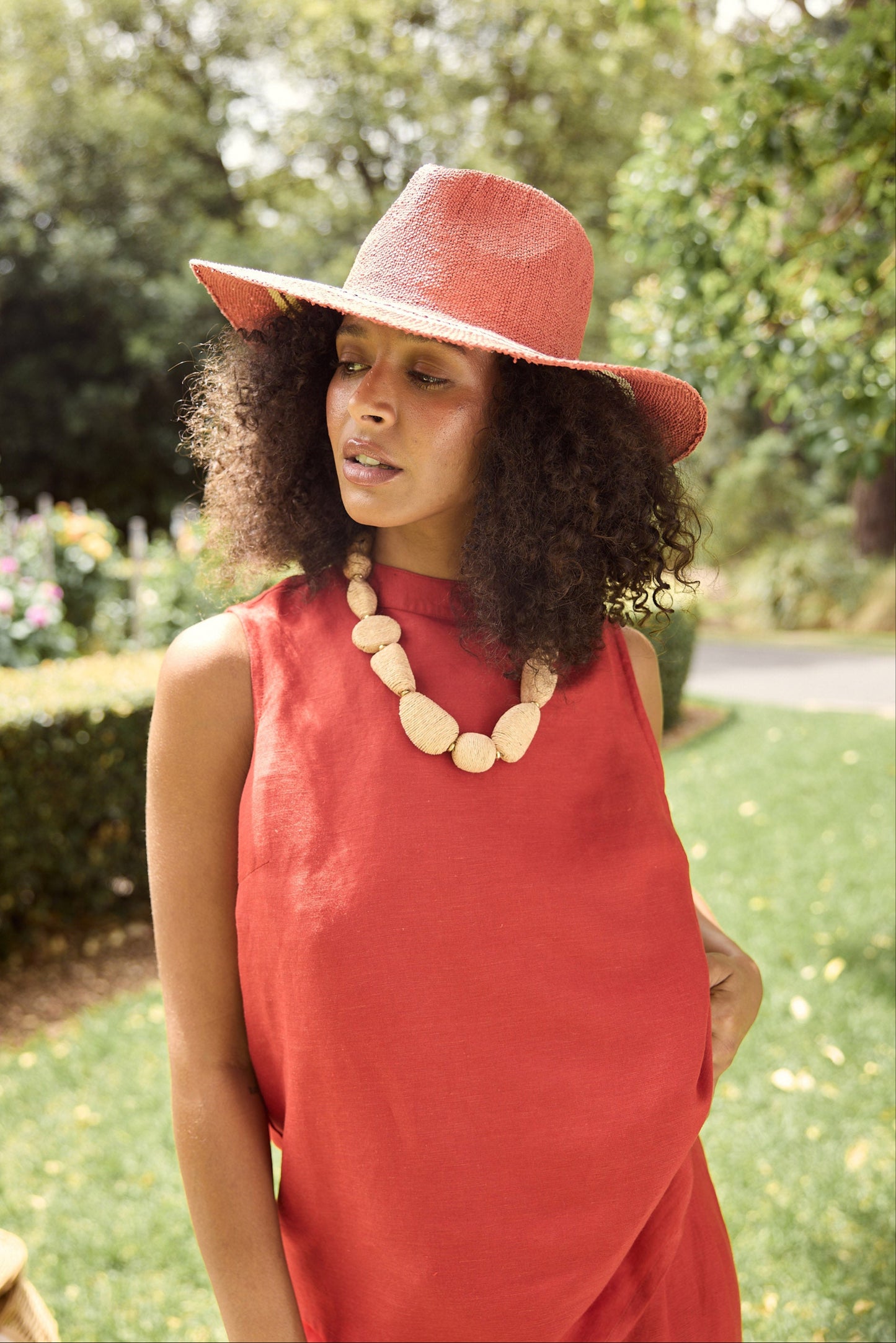 Woman wearing a red dress and hat with a necklace, standing outdoors.