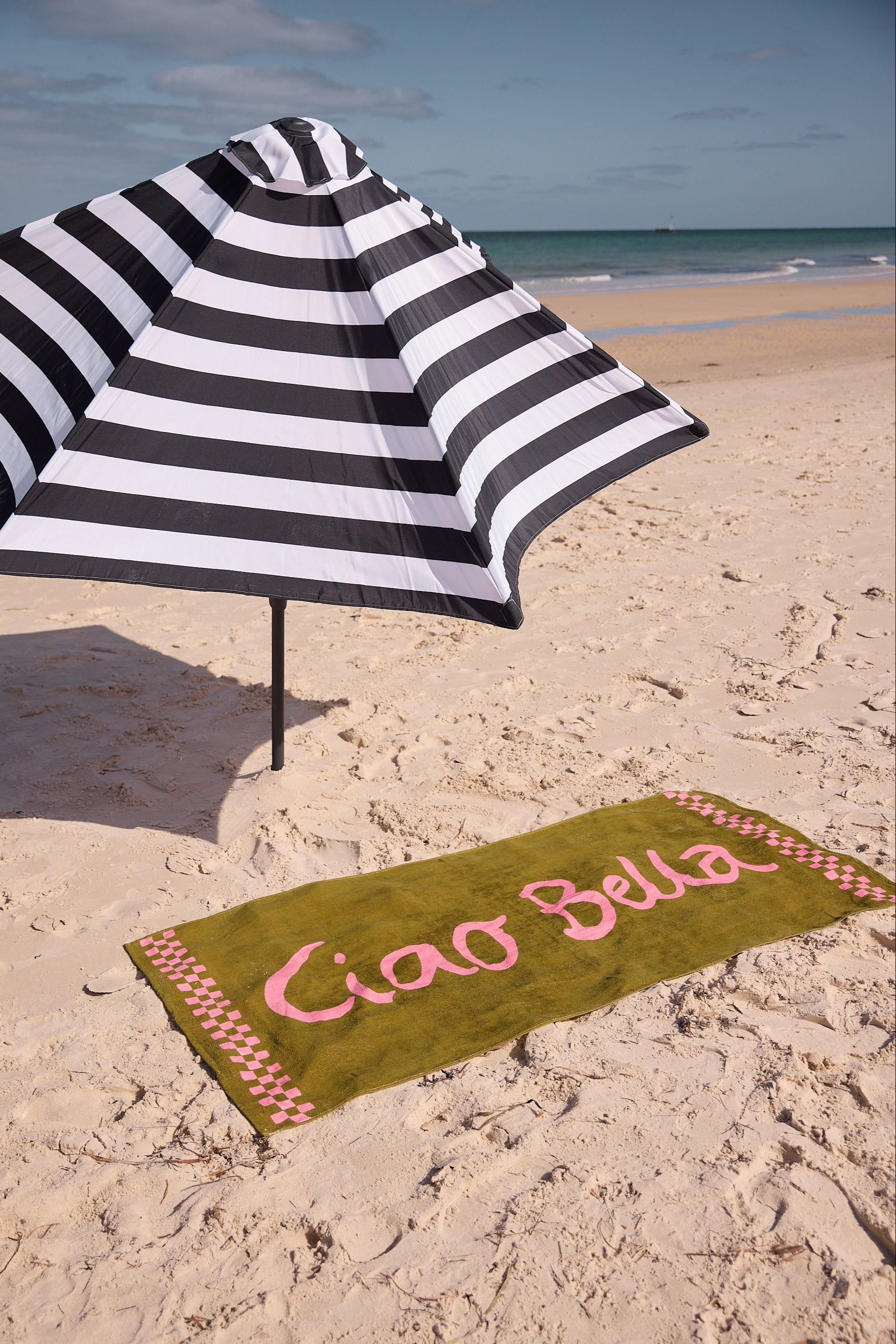 Beach scene with a black and white striped umbrella and a green towel with 'Ciao Bella' text on sand.
