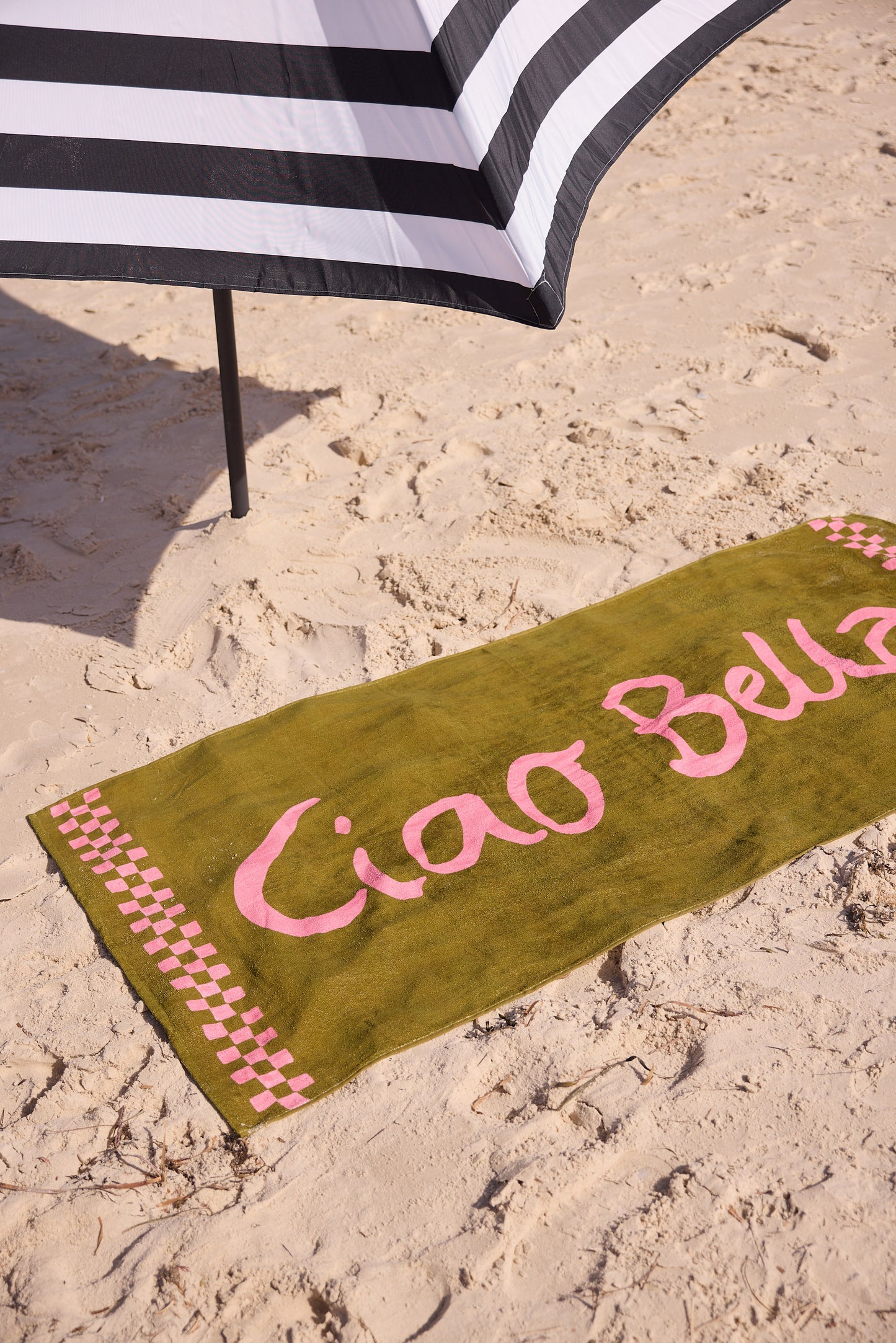 Green beach towel with 'Ciao Bella' text on sand, next to a striped umbrella