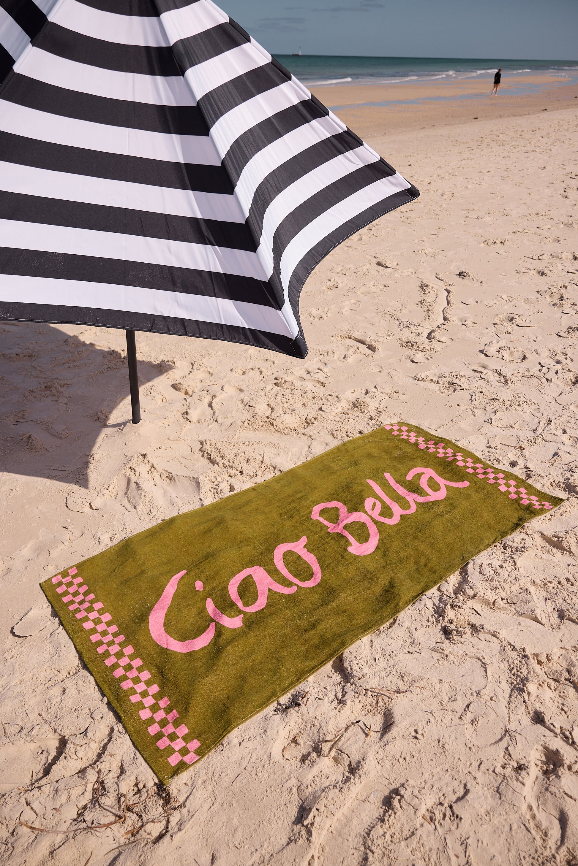 Beach towel with 'Ciao Bella' text on sand next to a striped umbrella