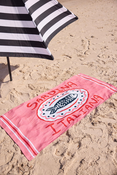 Pink beach towel with 'Sardegna Italiana' text and fish design on sand, next to a black and white striped umbrella.