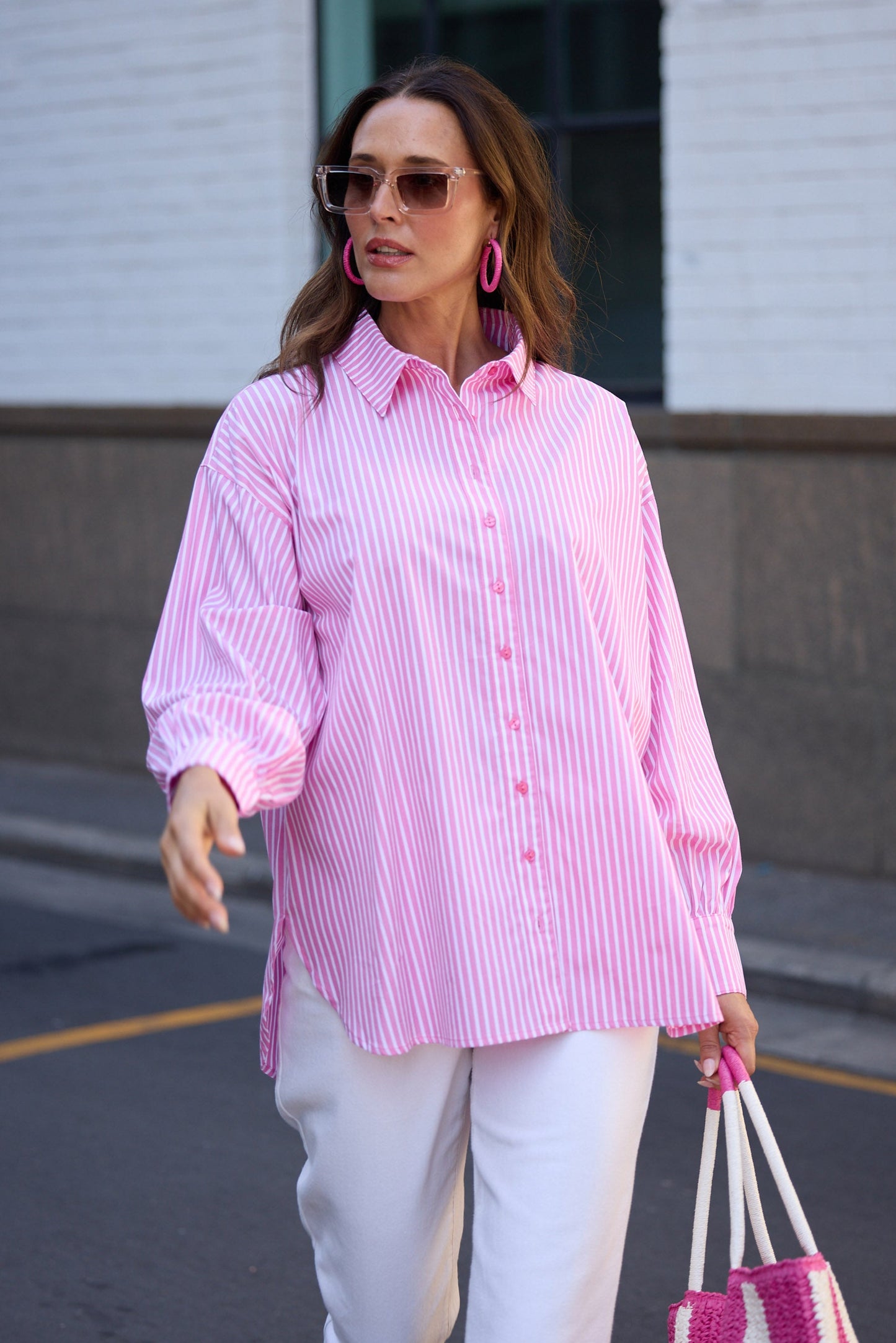 Woman wearing a pink striped shirt and white pants, holding a pink and white bag, walking on a street.