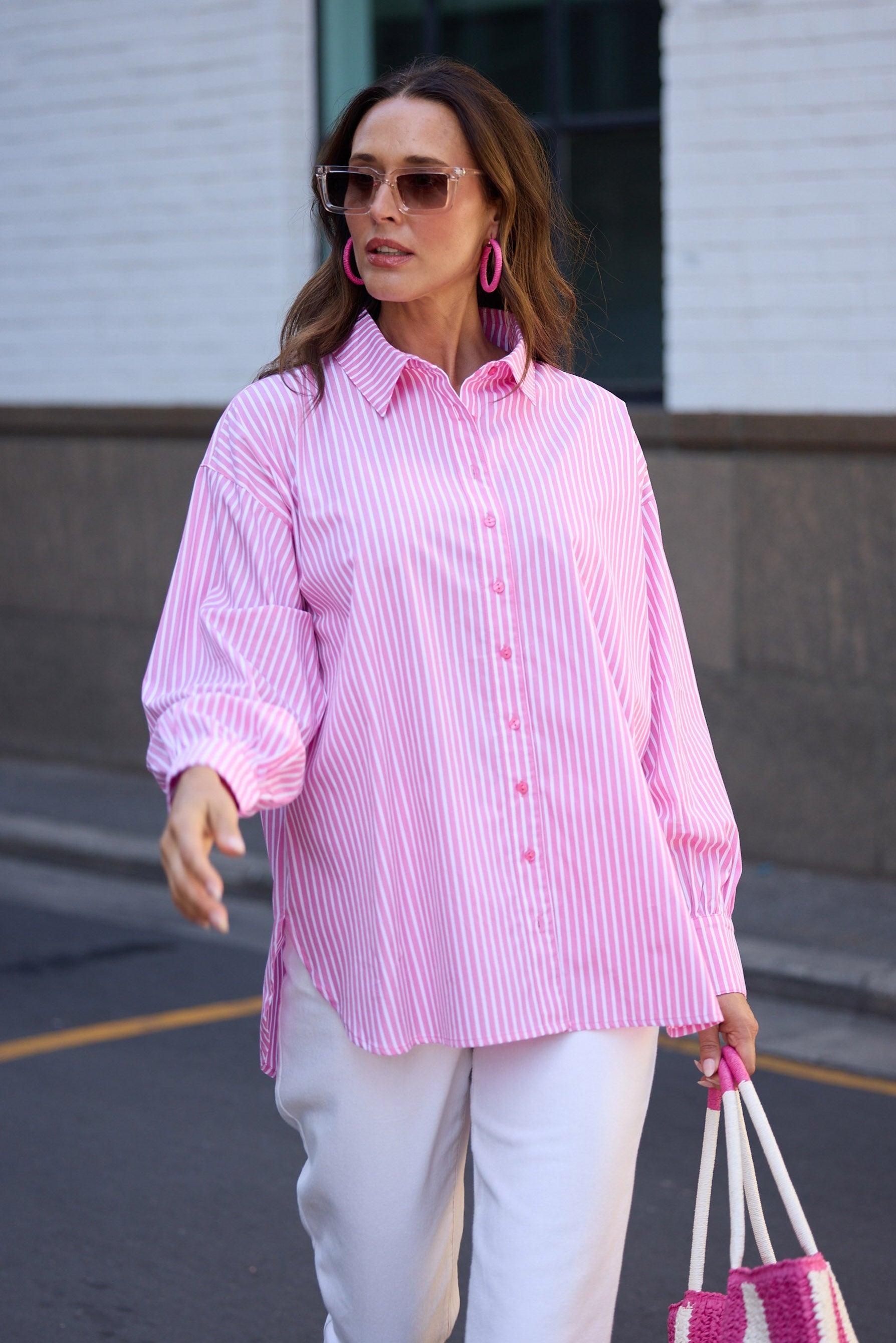 Woman wearing a pink striped shirt and white pants, holding a pink and white bag, walking on a street.