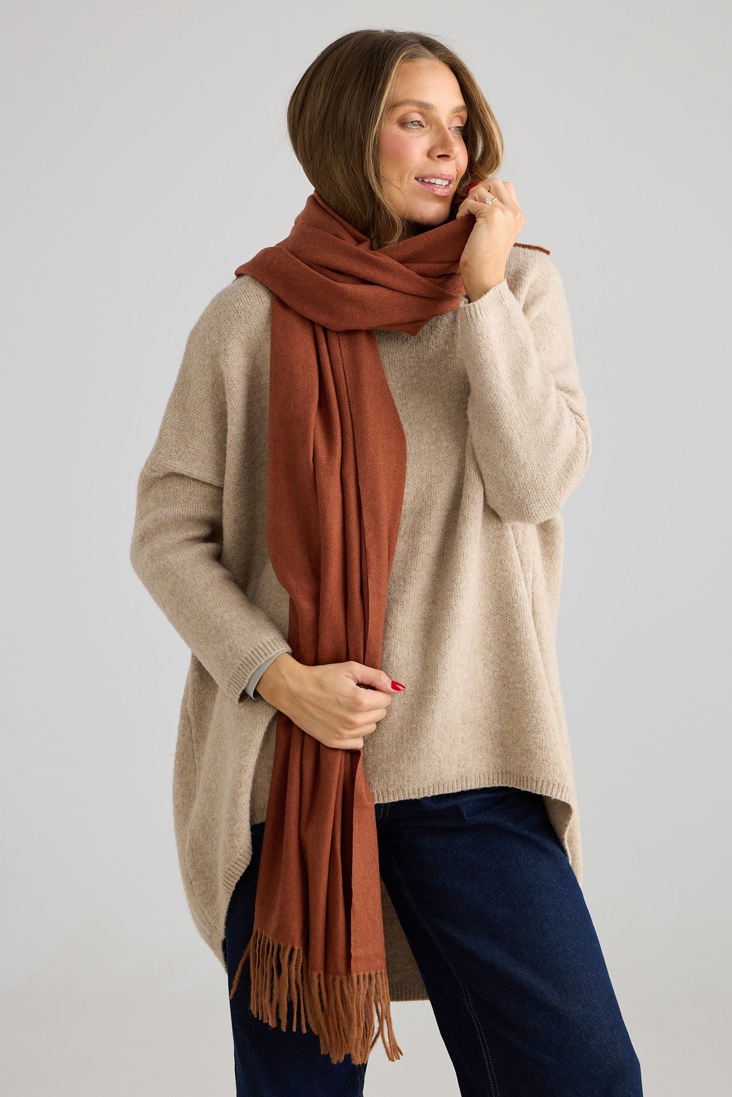 Woman wearing a beige sweater and blue pants with a rust-colored scarf on a plain background