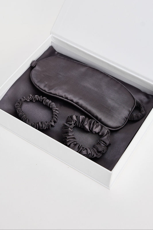 Dark gray eye mask and scrunchie set in an open white box on a light gray background