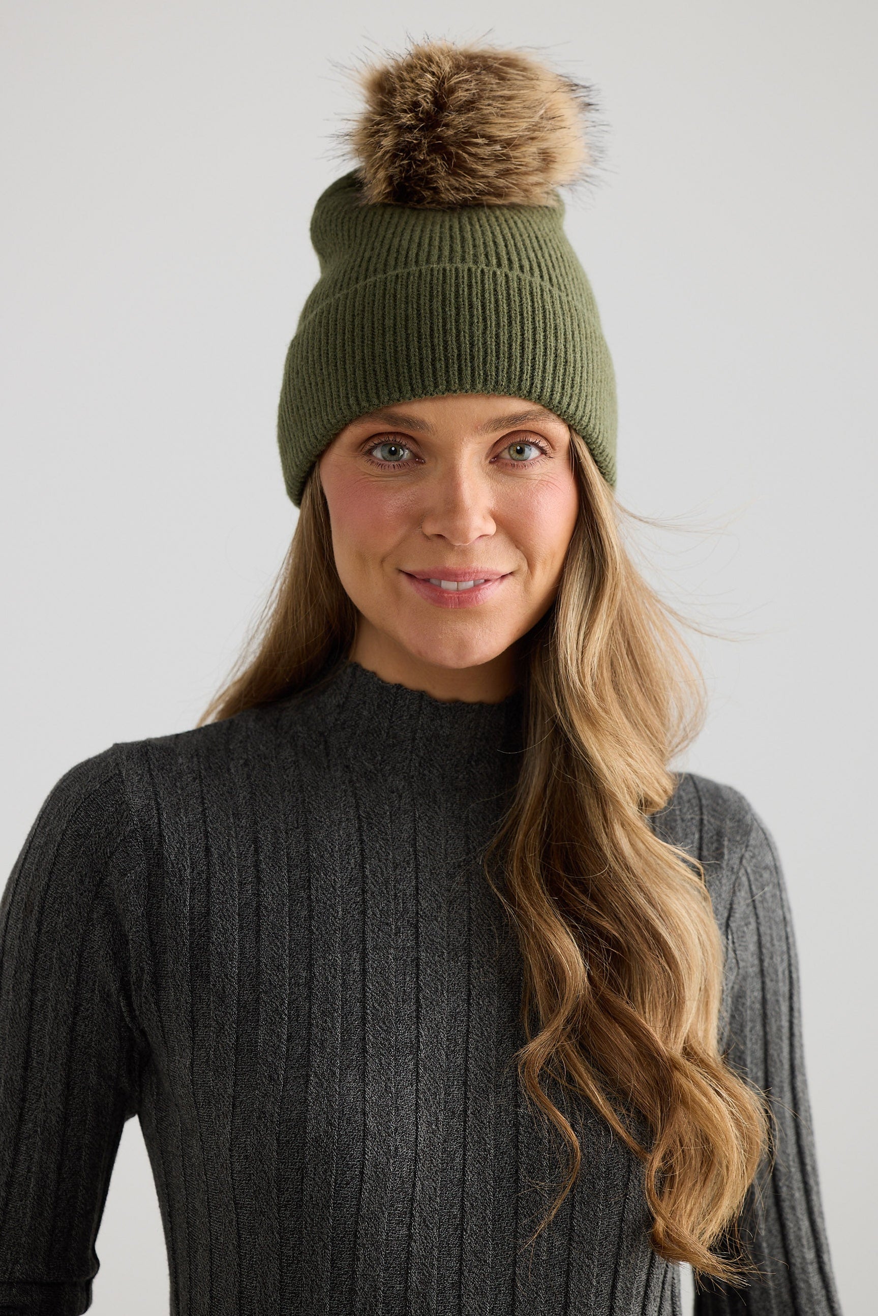 Woman wearing a green knit beanie with a fur pom-pom on a plain background