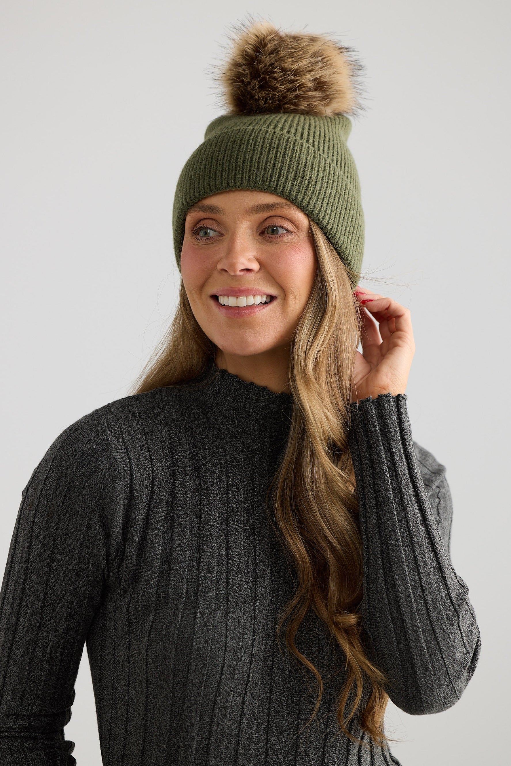 Woman wearing a green knit beanie with a fur pom-pom against a plain background