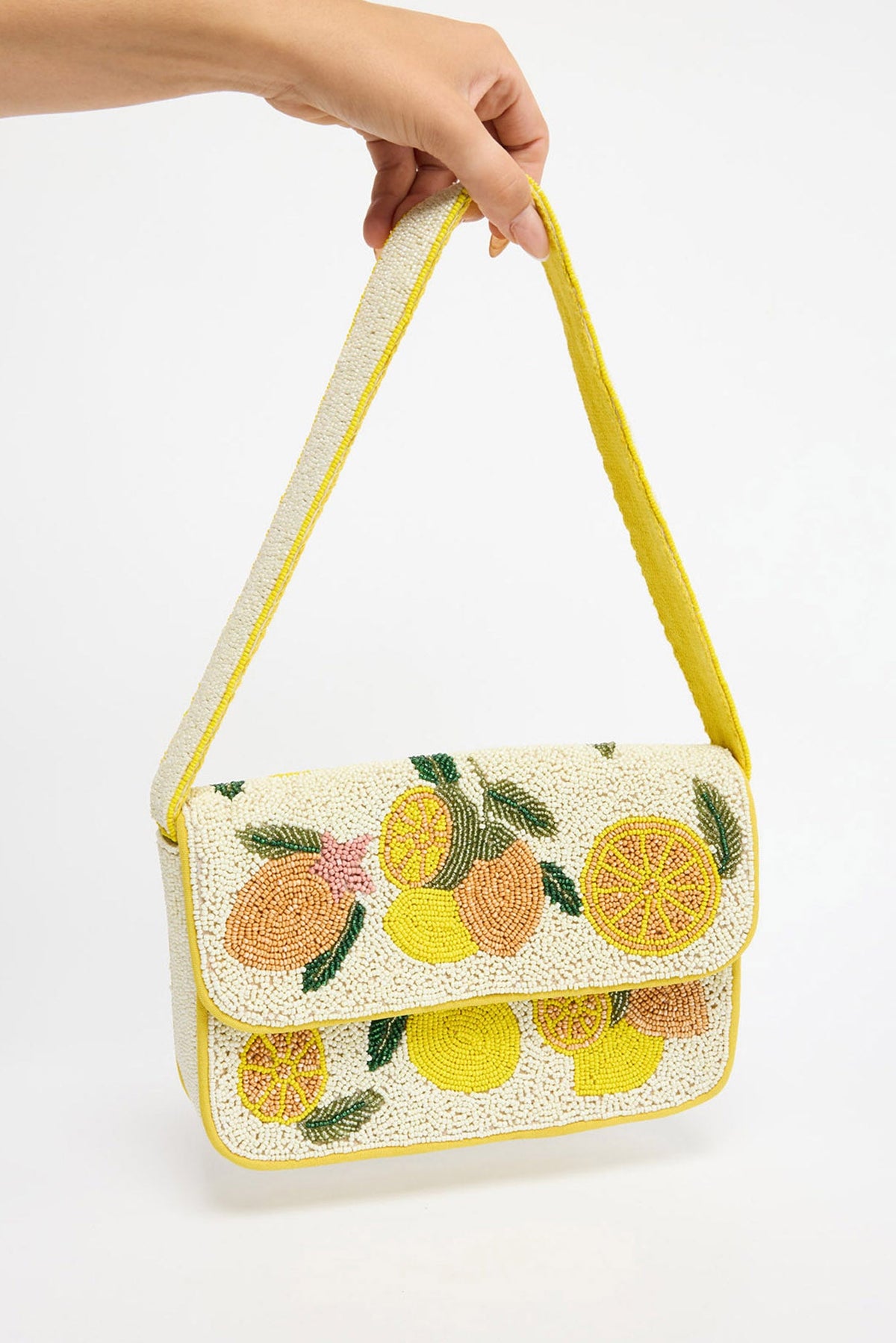 Rectangular structured shoulder bag fully covered in cream seed beads with a bold beaded lemon and citrus slice motif in yellow, orange and green, yellow trim and a long beaded strap, held by hand against a white background
