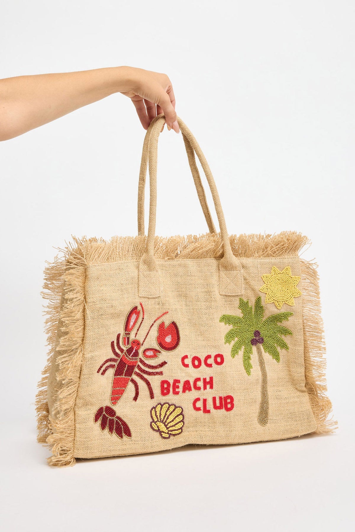 Large natural jute tote bag with fringe trim along the sides, beaded Coco Beach Club lettering and tropical motifs including a red lobster, green palm tree, yellow sun and shell, held by hand against a white background