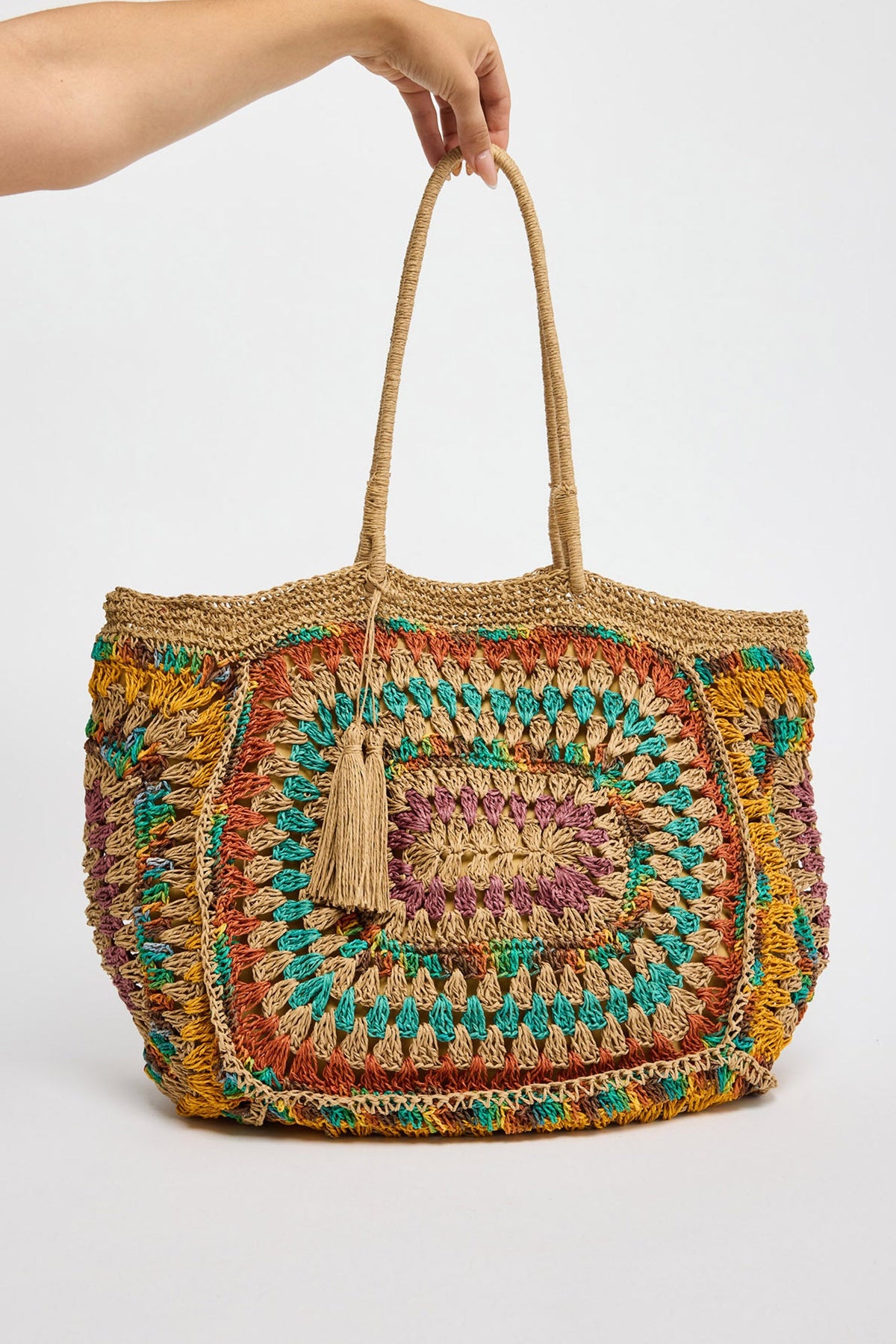 Large handwoven crochet tote in natural raffia with a large multicolour granny square panel in teal, terracotta, mustard and lilac tones, a tassel zip pull and twisted rope handles, held by hand against a white background