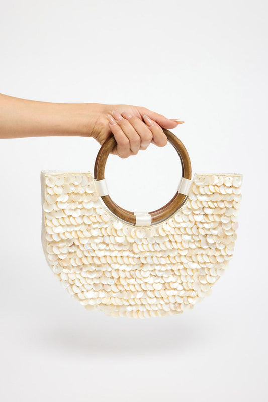 Half-moon shaped bag fully covered in overlapping cream shell discs with a warm iridescent sheen, held by a round tortoiseshell-tone acrylic handle against a white background