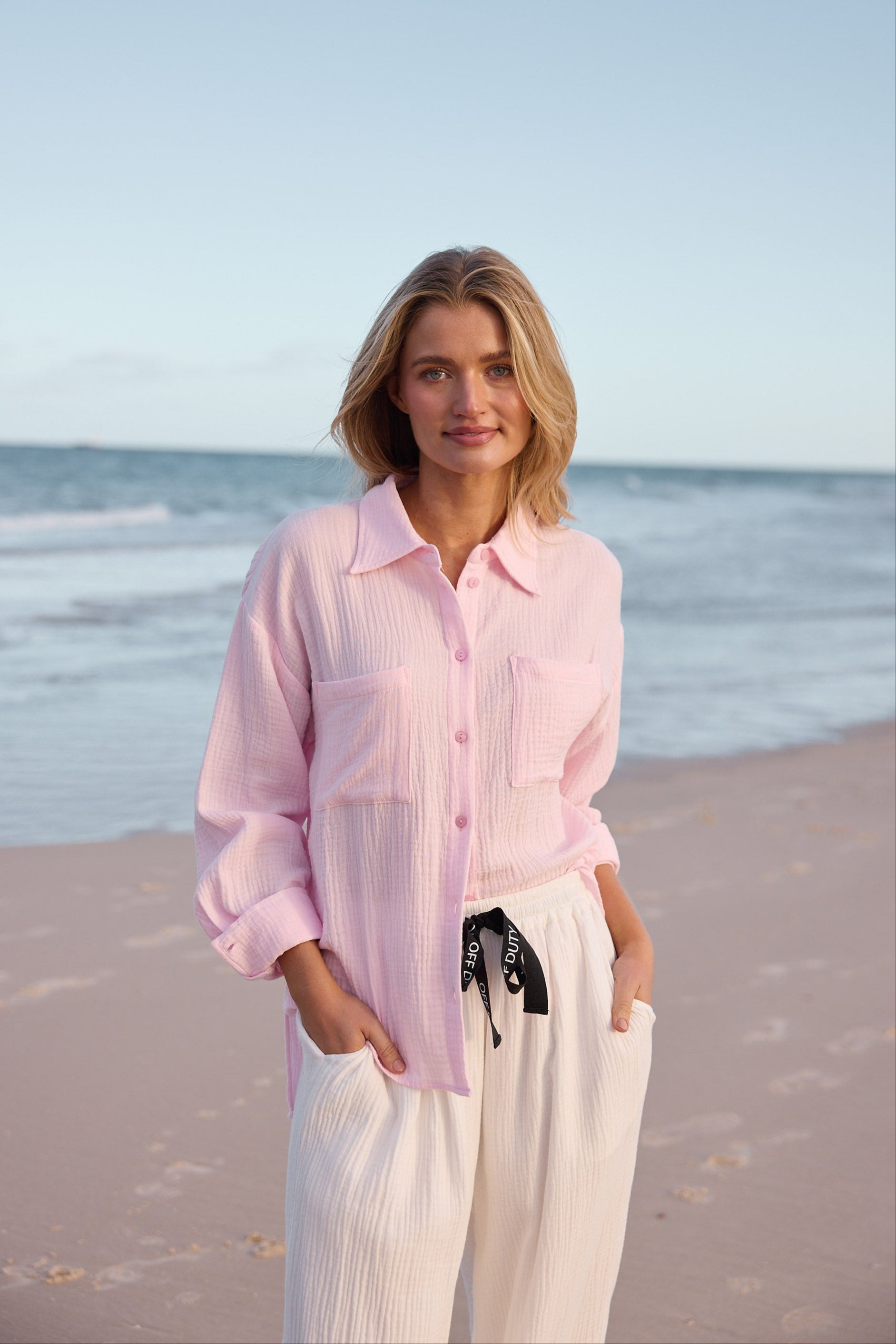 Woman in a pink shirt and white pants standing on a beach with ocean in the background