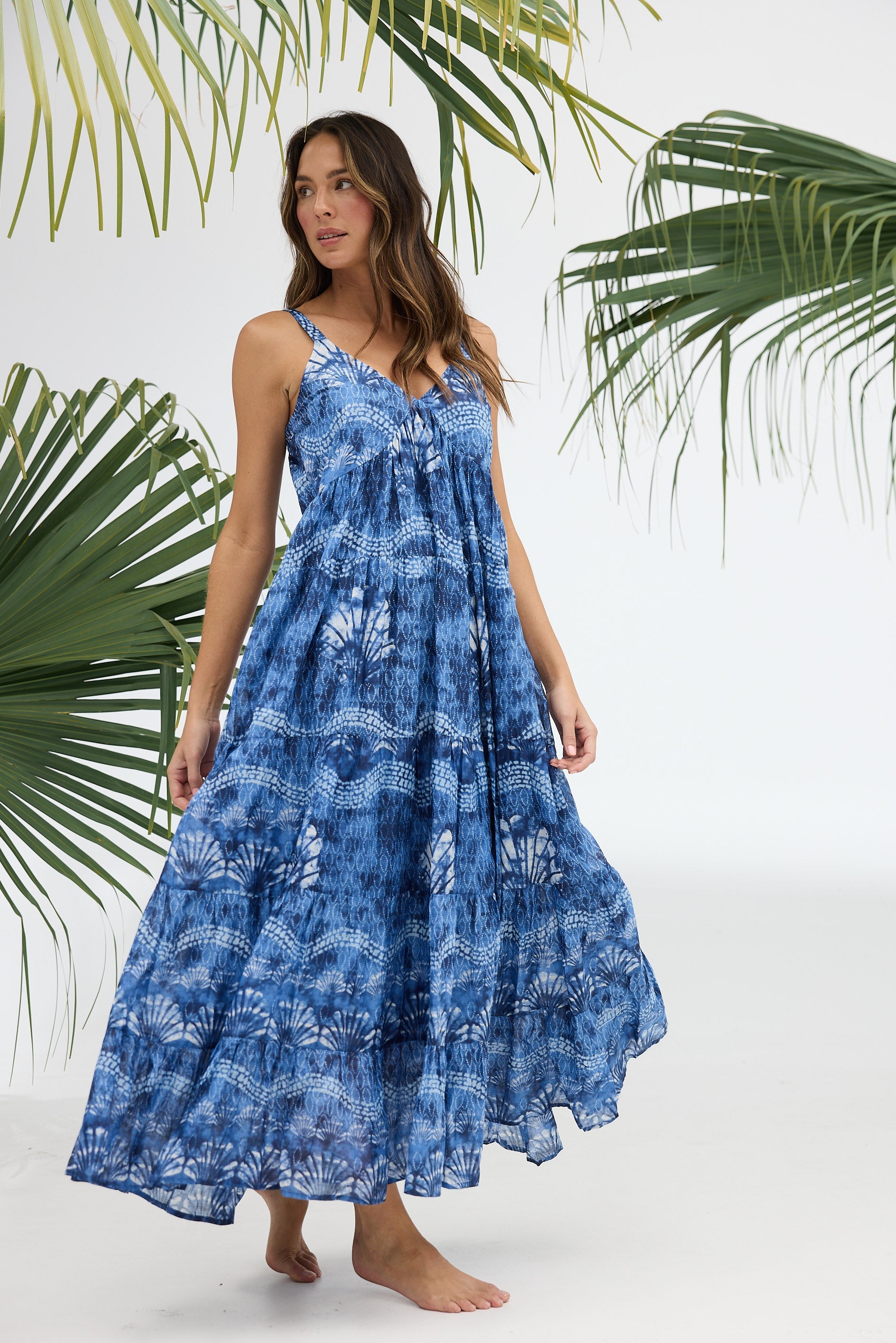 Woman wearing a blue floral dress standing against a white background with green palm leaves.