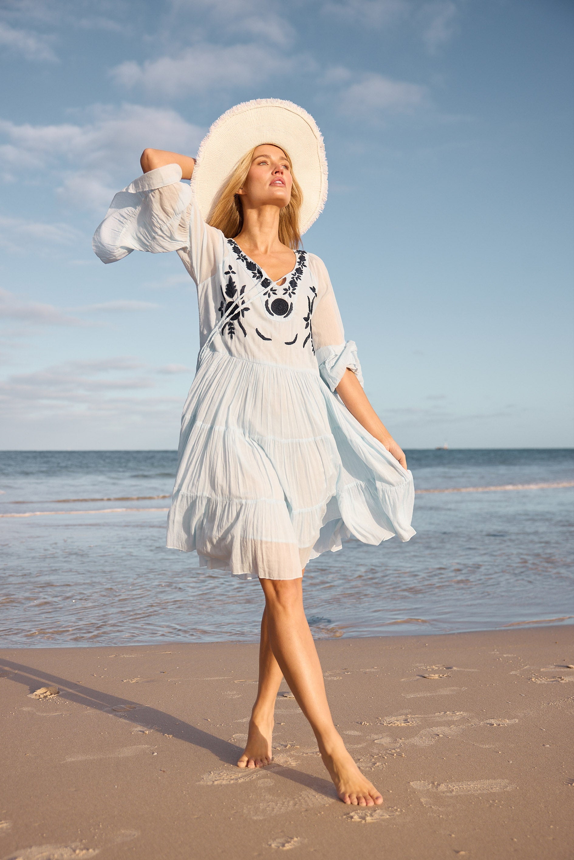 Woman in a white dress and hat walking on a beach with ocean and sky in the background