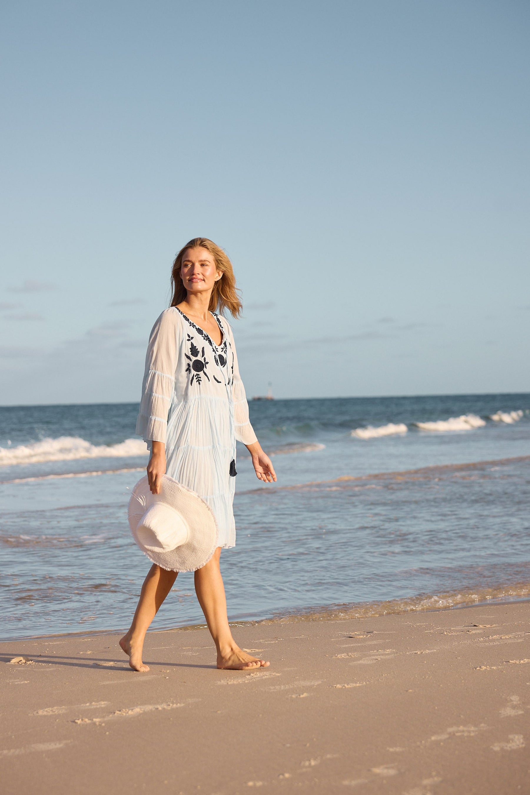 Woman walking on a beach with a clear blue sky and ocean waves in the background.