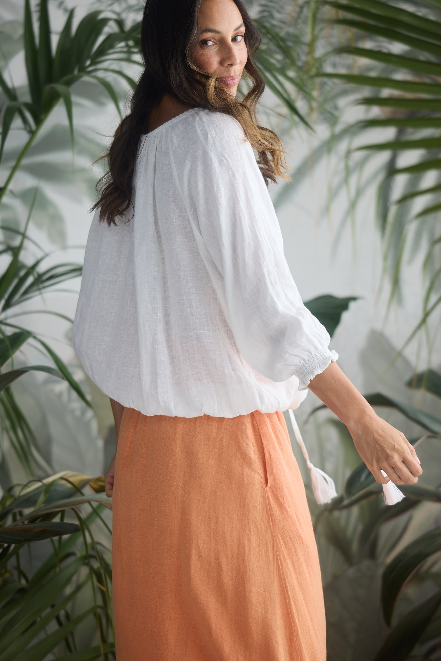 Woman wearing a white blouse and orange skirt standing among green plants.