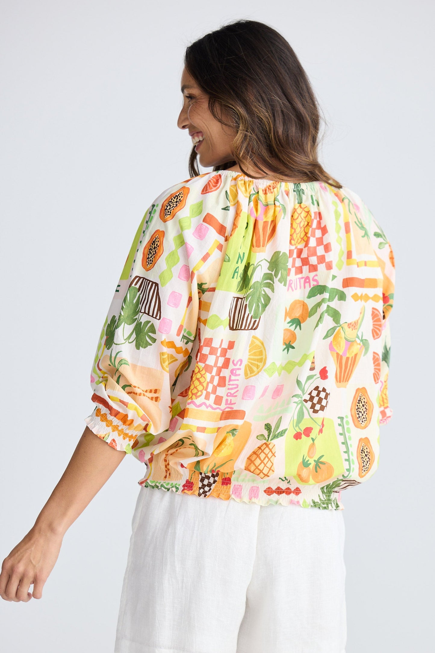 Woman wearing a colorful fruit print blouse and white shorts on a plain background