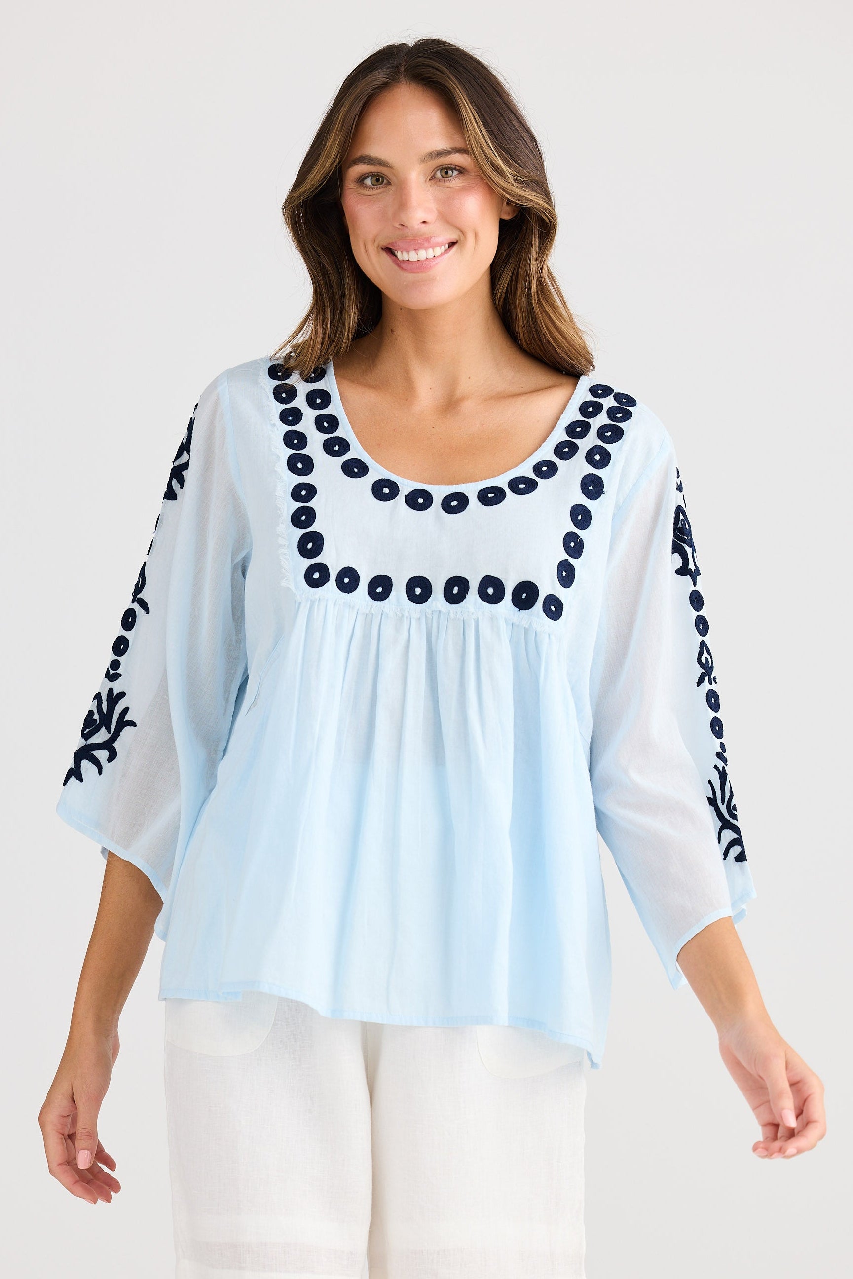 Woman wearing a light blue blouse with black embroidery and white shorts on a white background