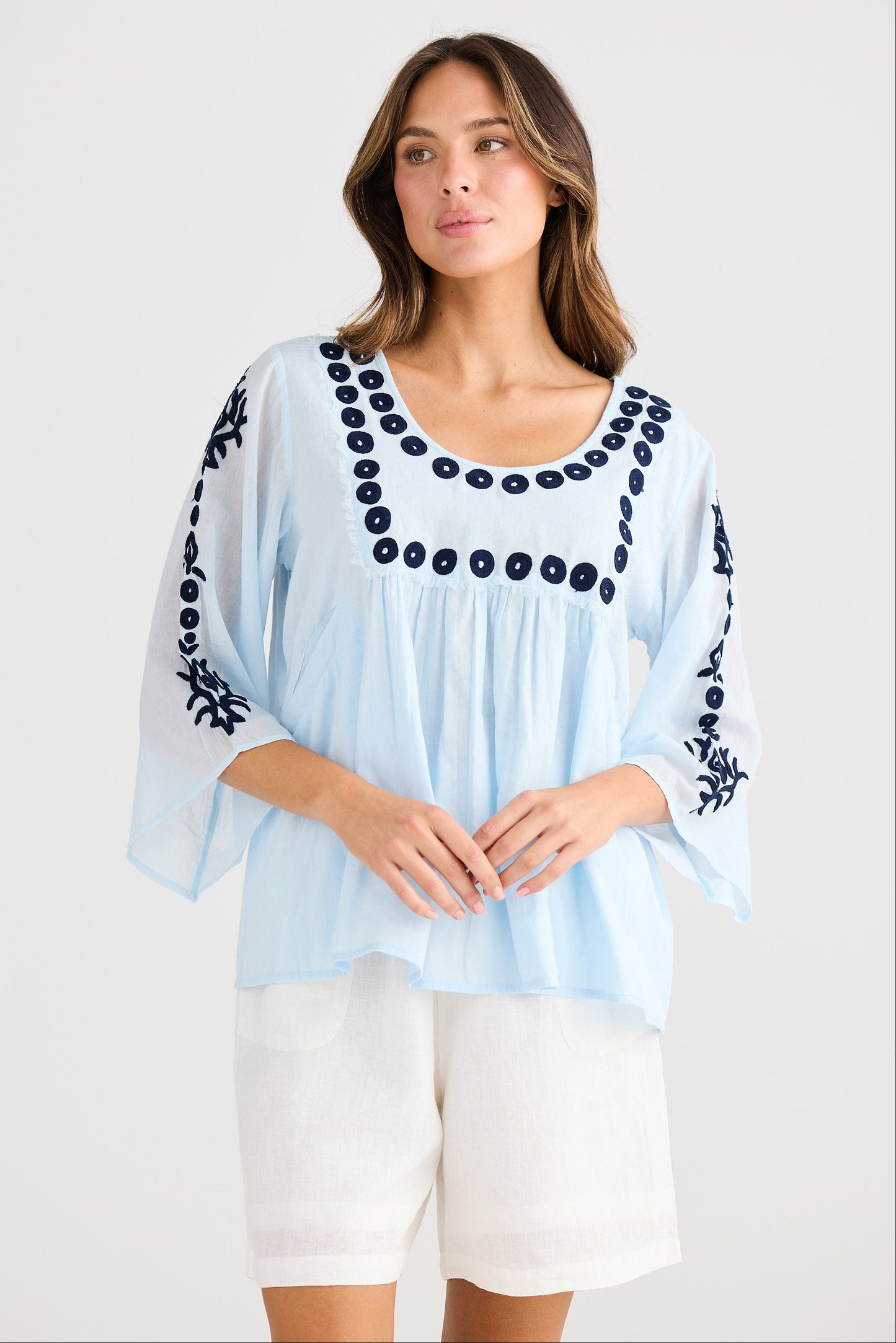 Woman wearing a light blue blouse with black embroidery and white shorts on a white background