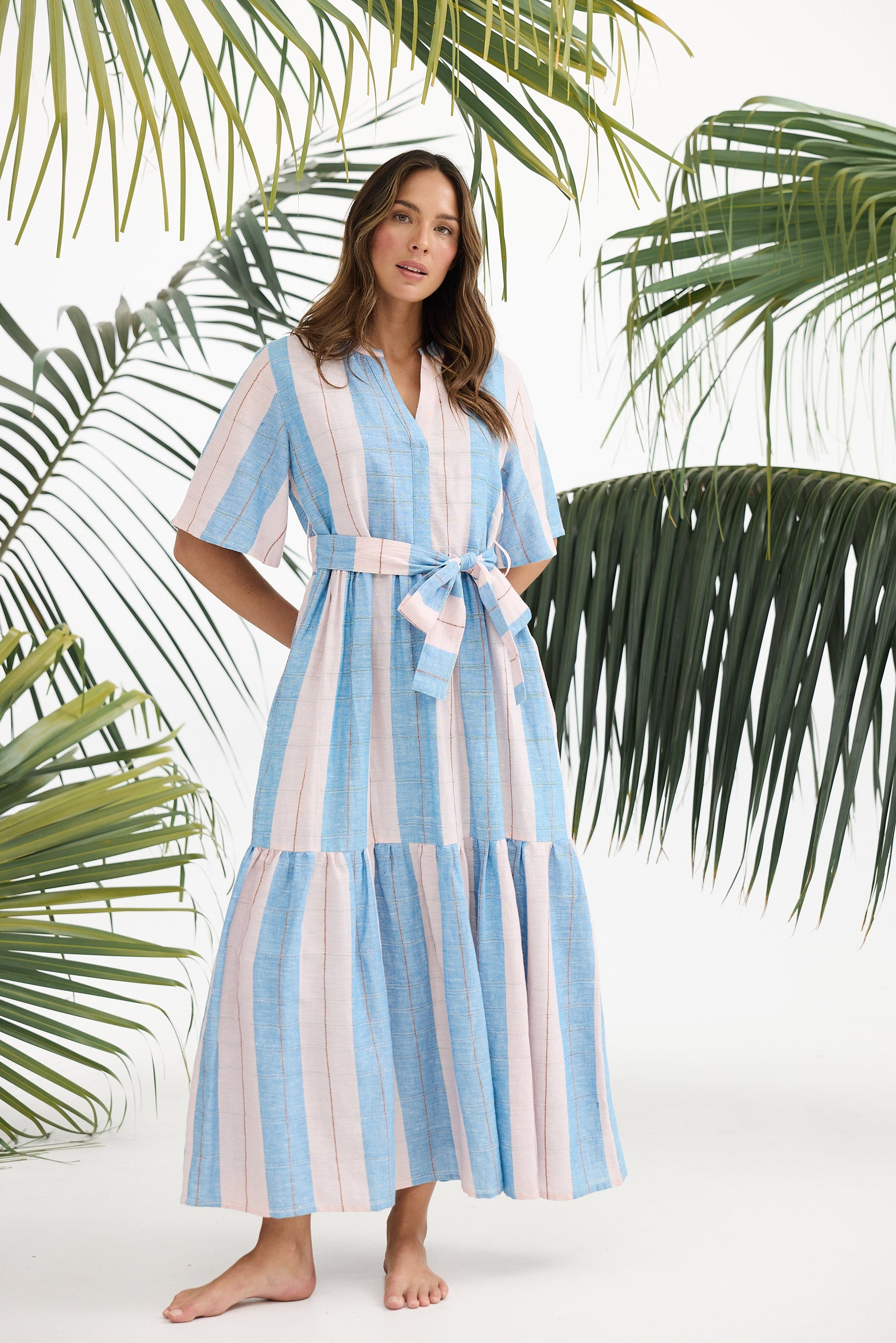 Woman wearing a blue and white striped dress with a belt, standing against a white background with palm leaves.