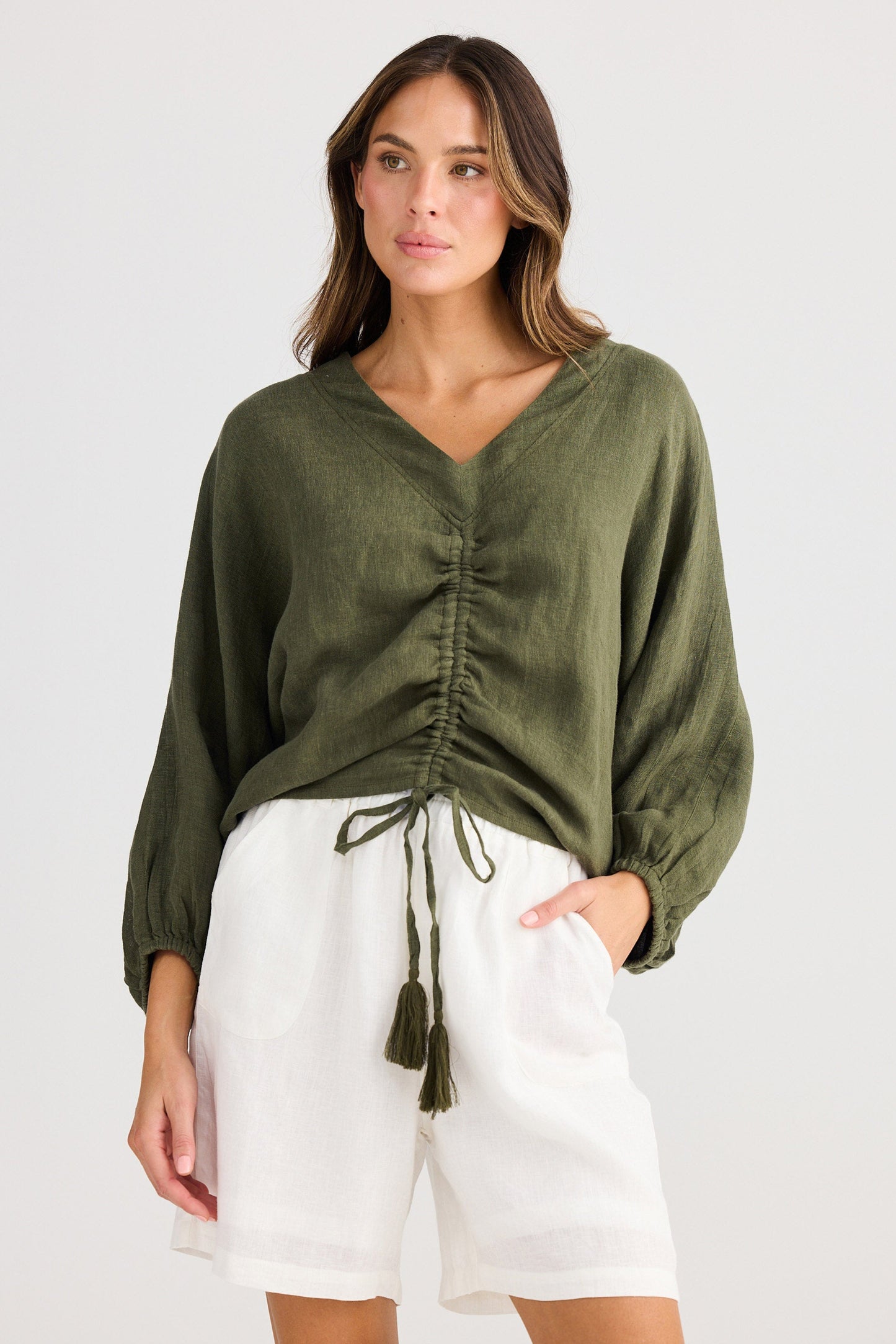 Woman wearing an olive green top with a tie front and white shorts on a white background