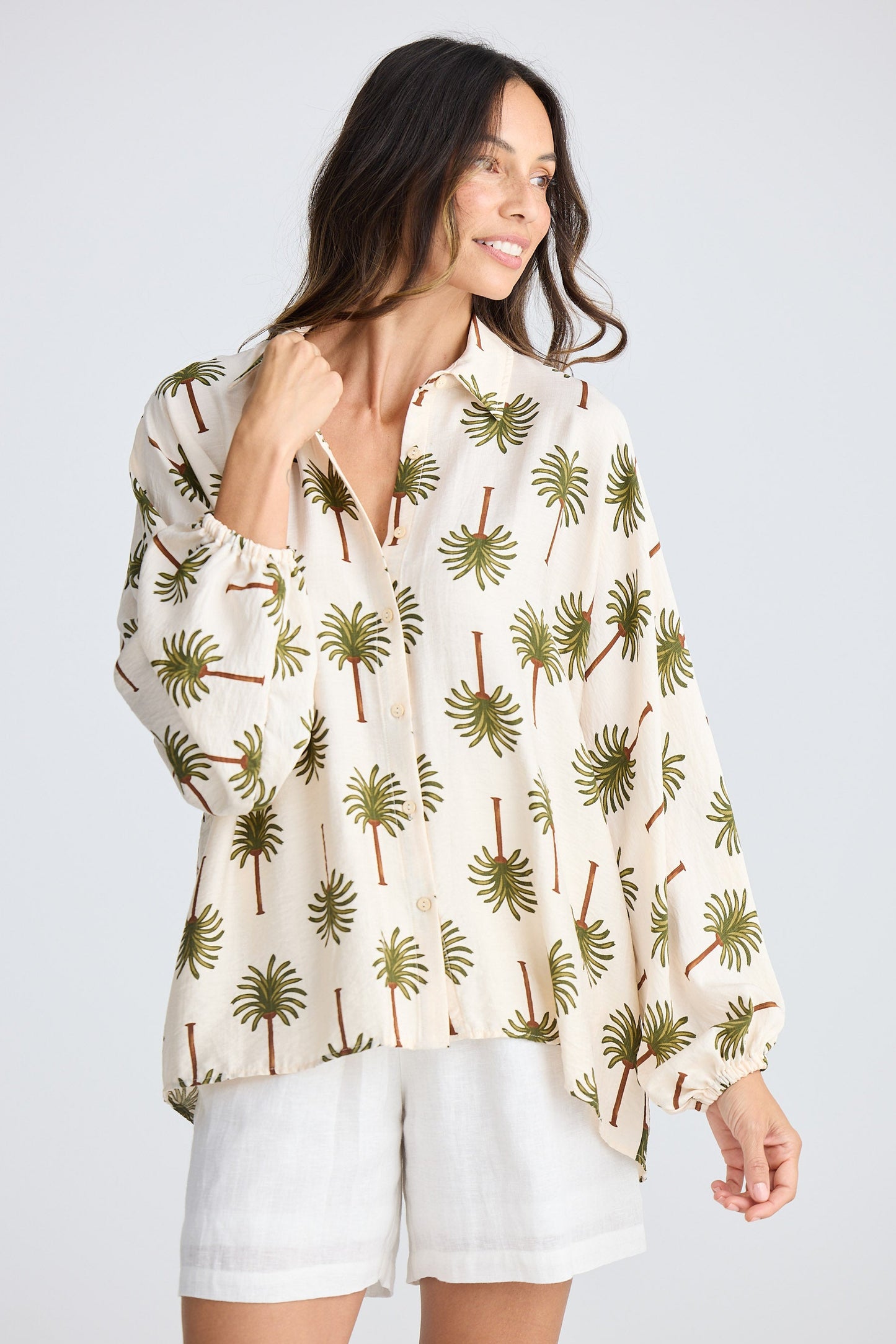 Woman wearing a white blouse with palm tree pattern and white shorts on a light gray background