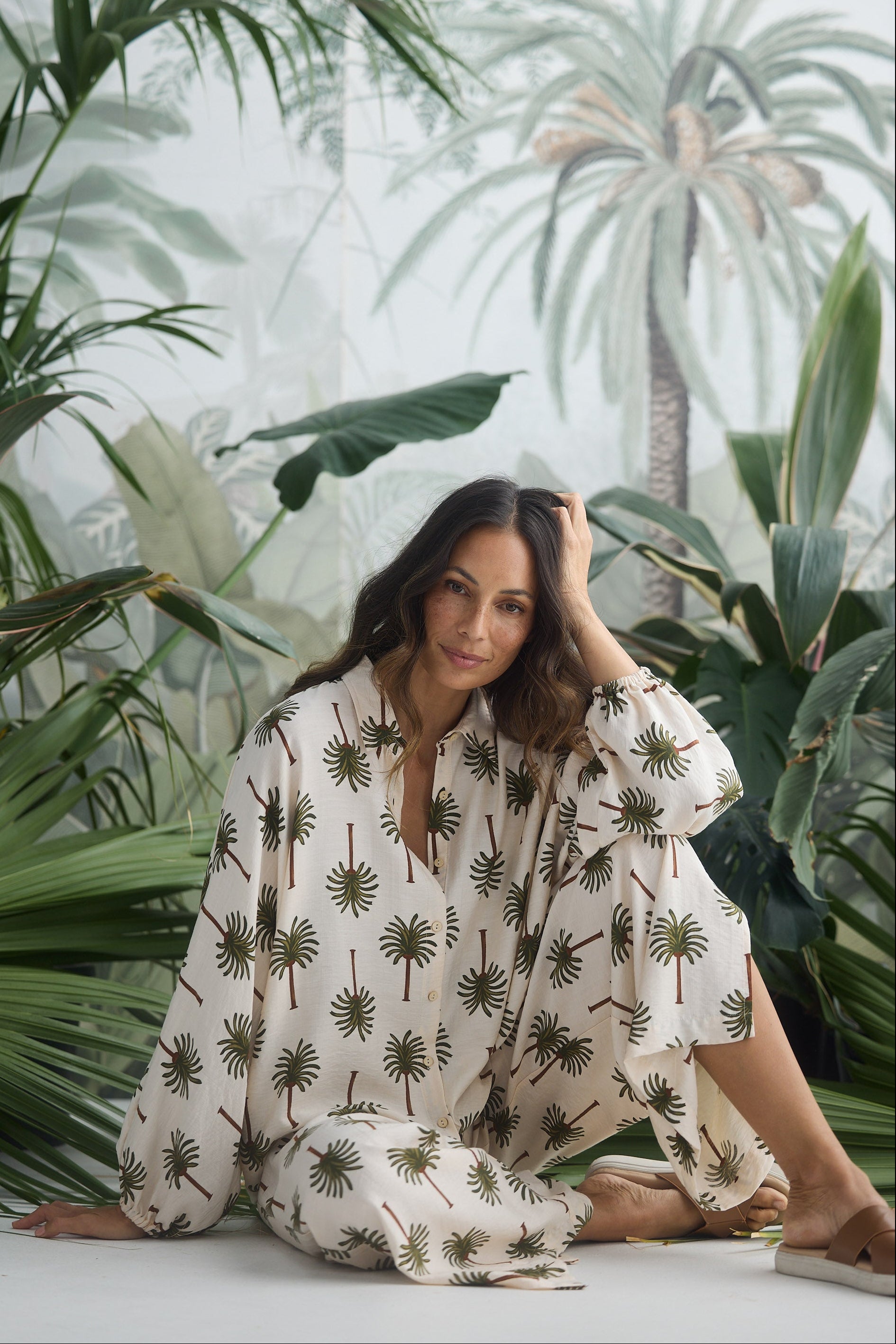 Woman in a white pants and shirt with palm tree pattern sitting among tropical plants